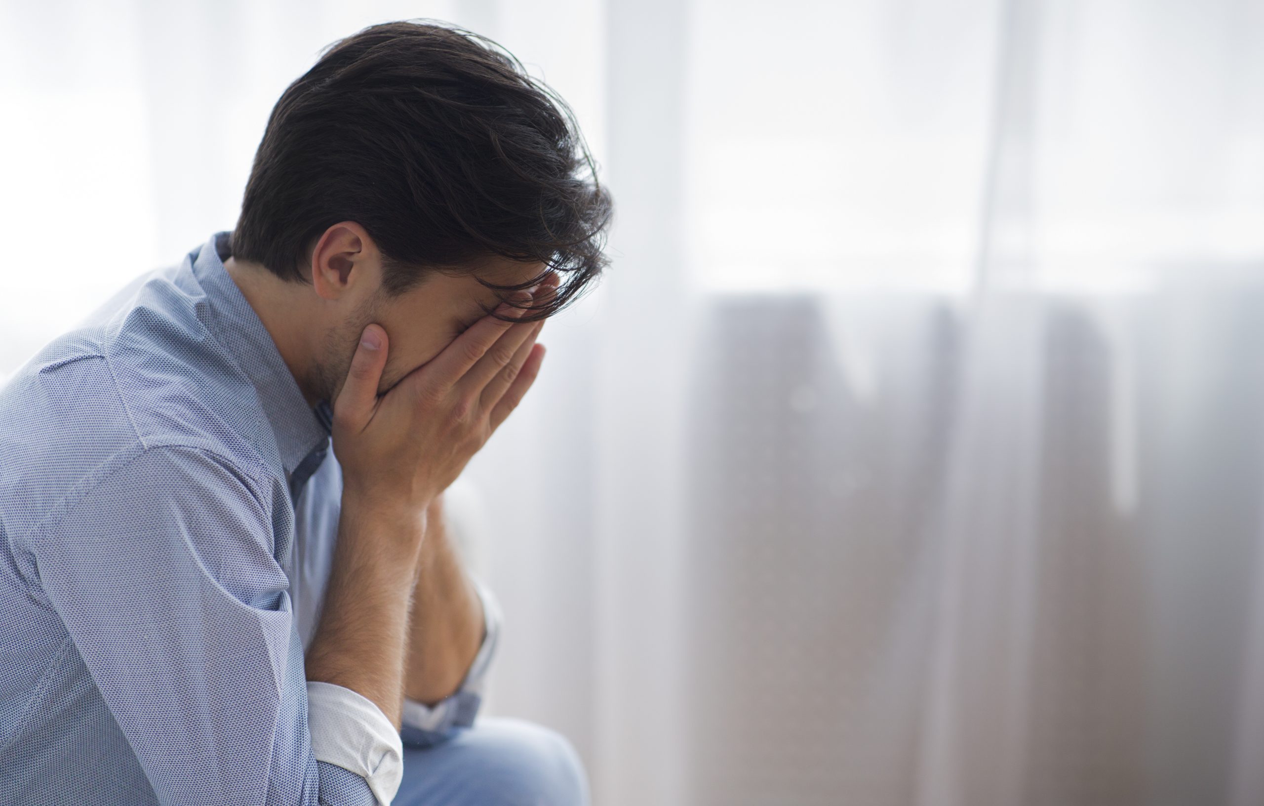 Male Depression. Unrecognizable Frustrated Man Covering Face With Hands Crying Sitting Against Window. Copy Space