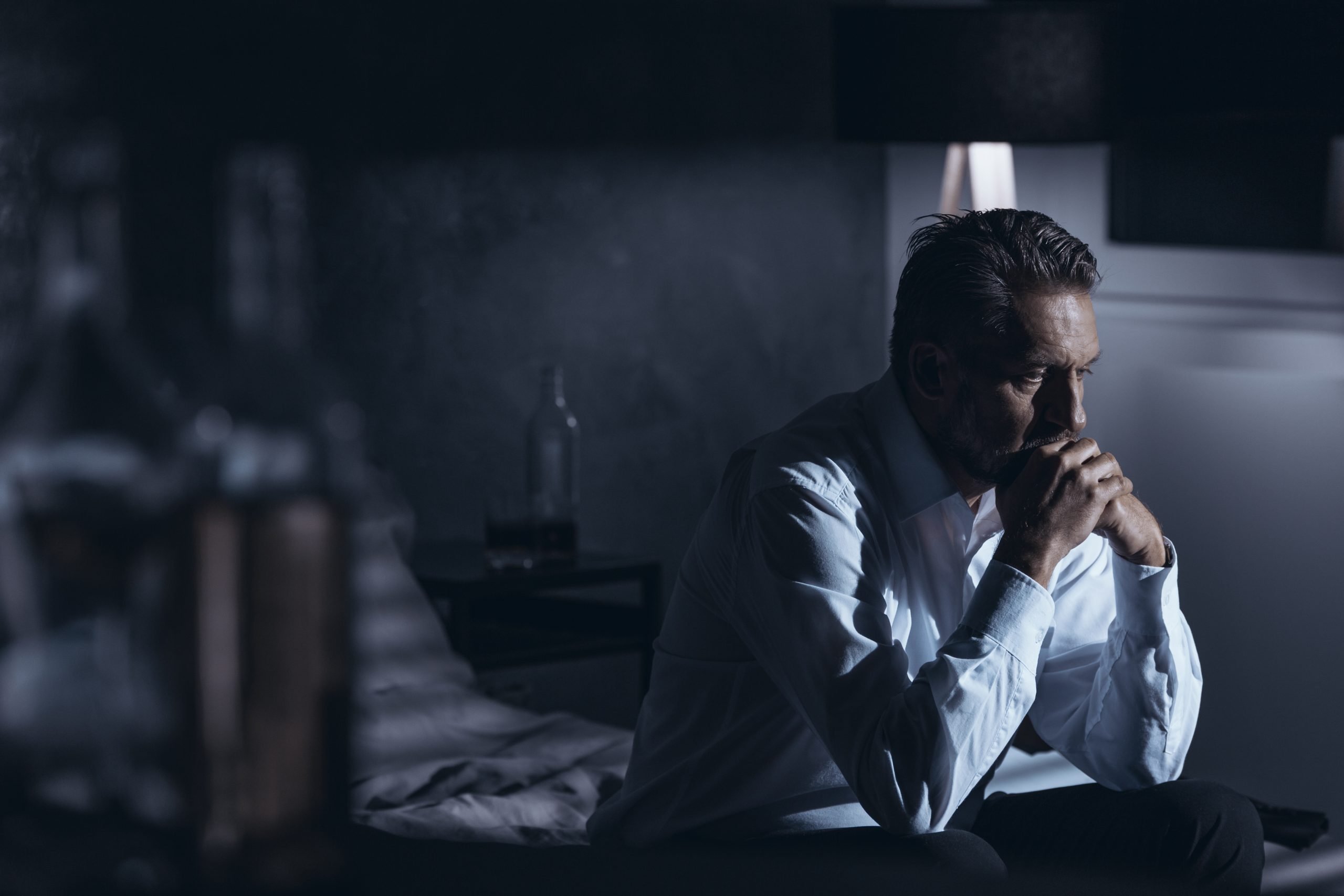 Portrait of a lonely mature man with depression sitting on a bed in a gray room with bottles of alcohol standing around