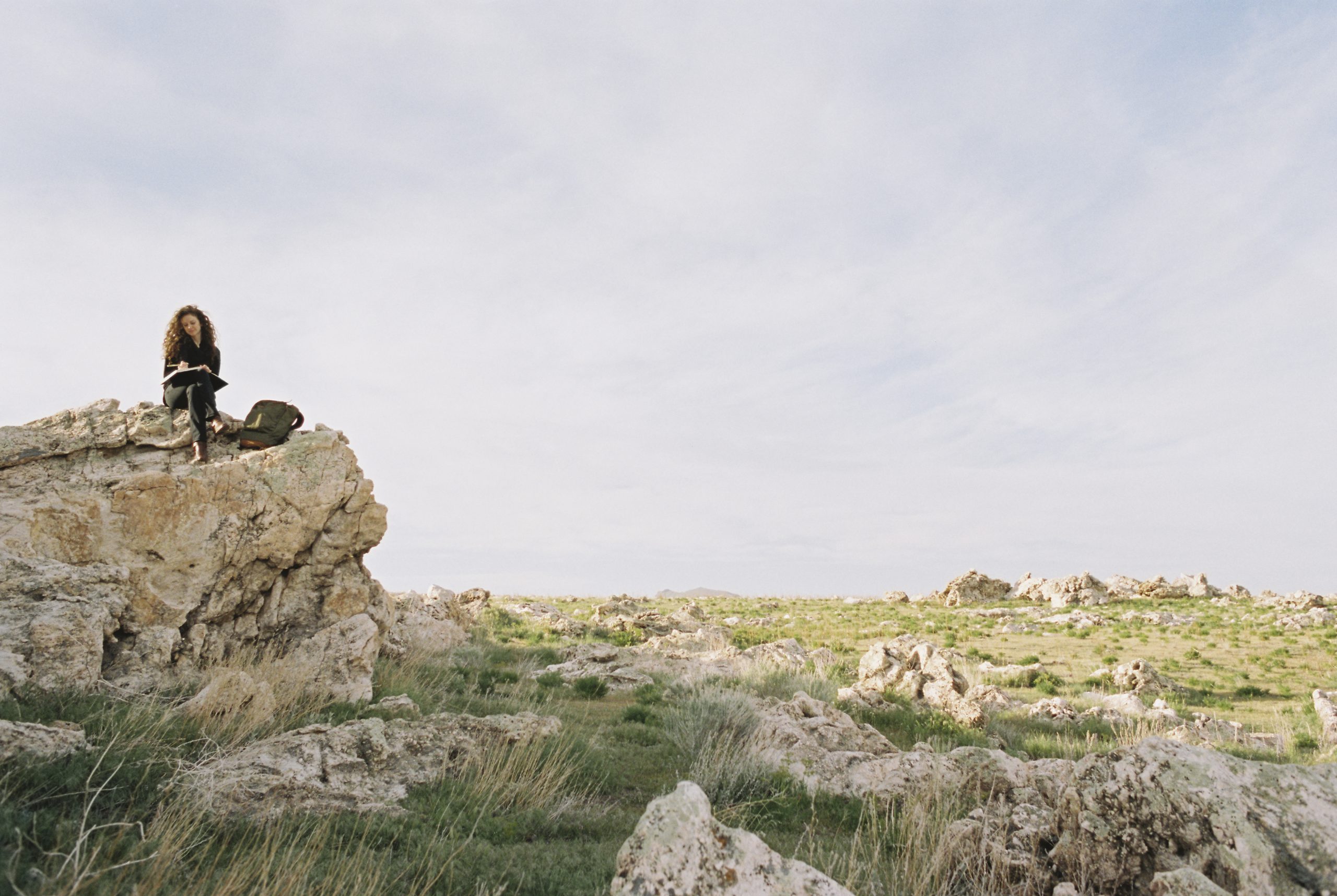 A woman sitting on a rock, writing in a notebook, a backpack lying by her feet.