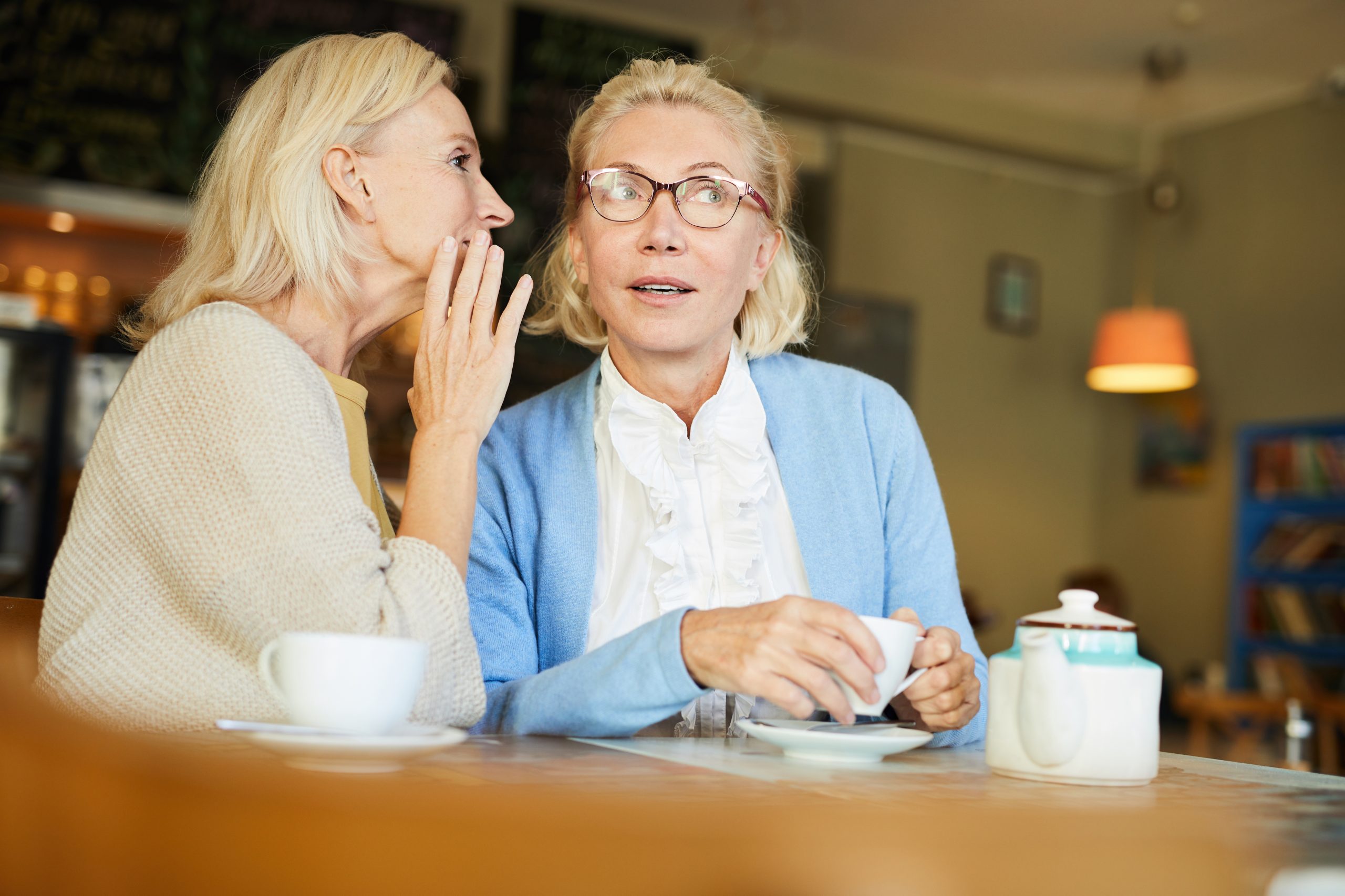 One of pretty aged females telling secret to her friend by cup of tea in cafe