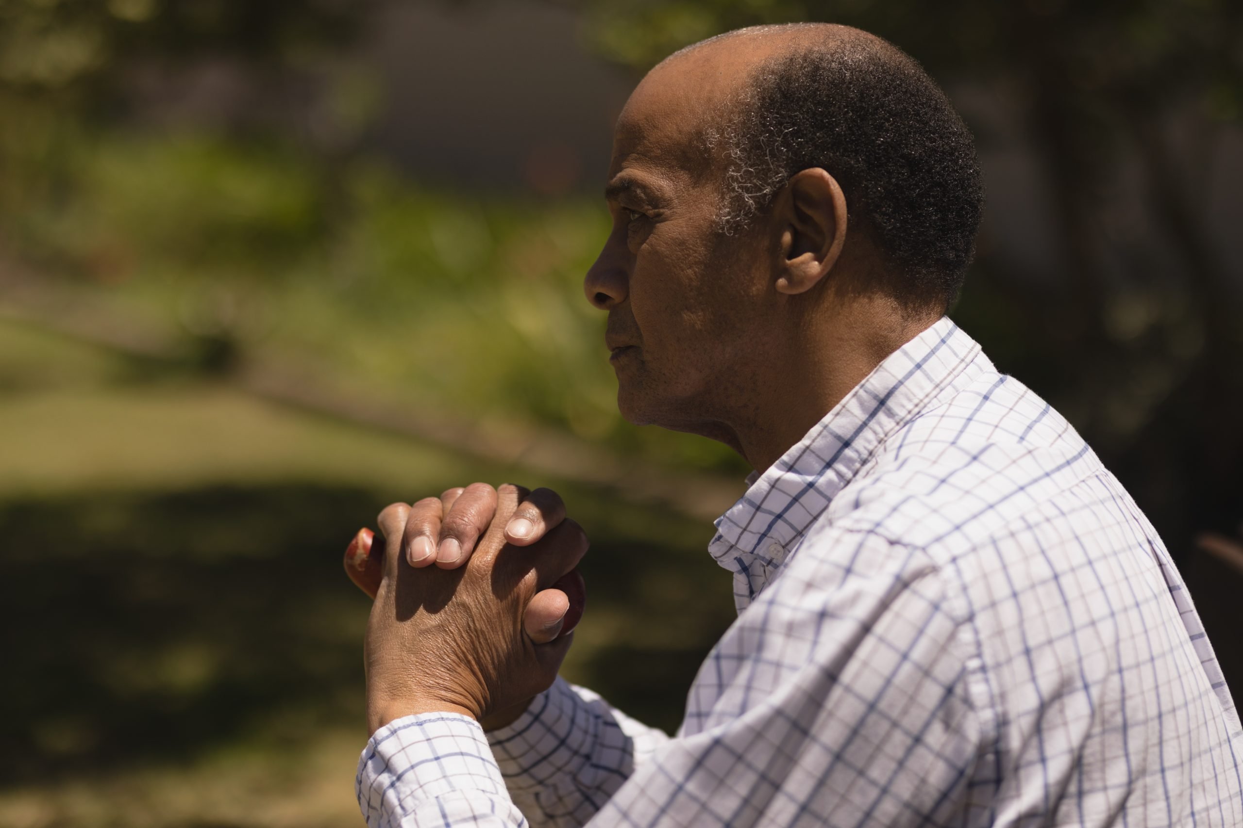 Side view of thoughtful senior man hands leaning on a cane while sitting on bench on sunny day in garden