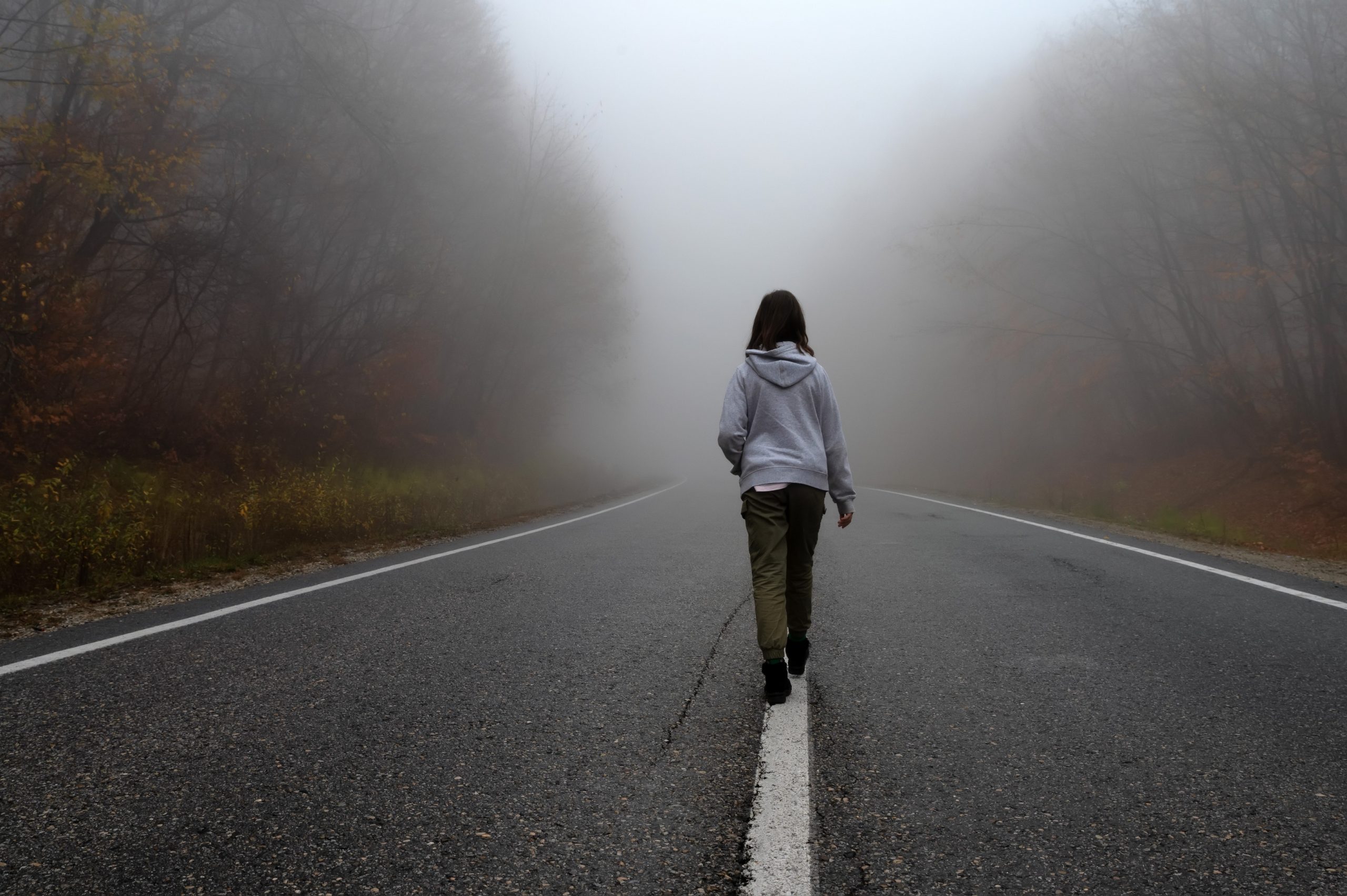 Back view of young woman walking in autumn forest up to foggy road