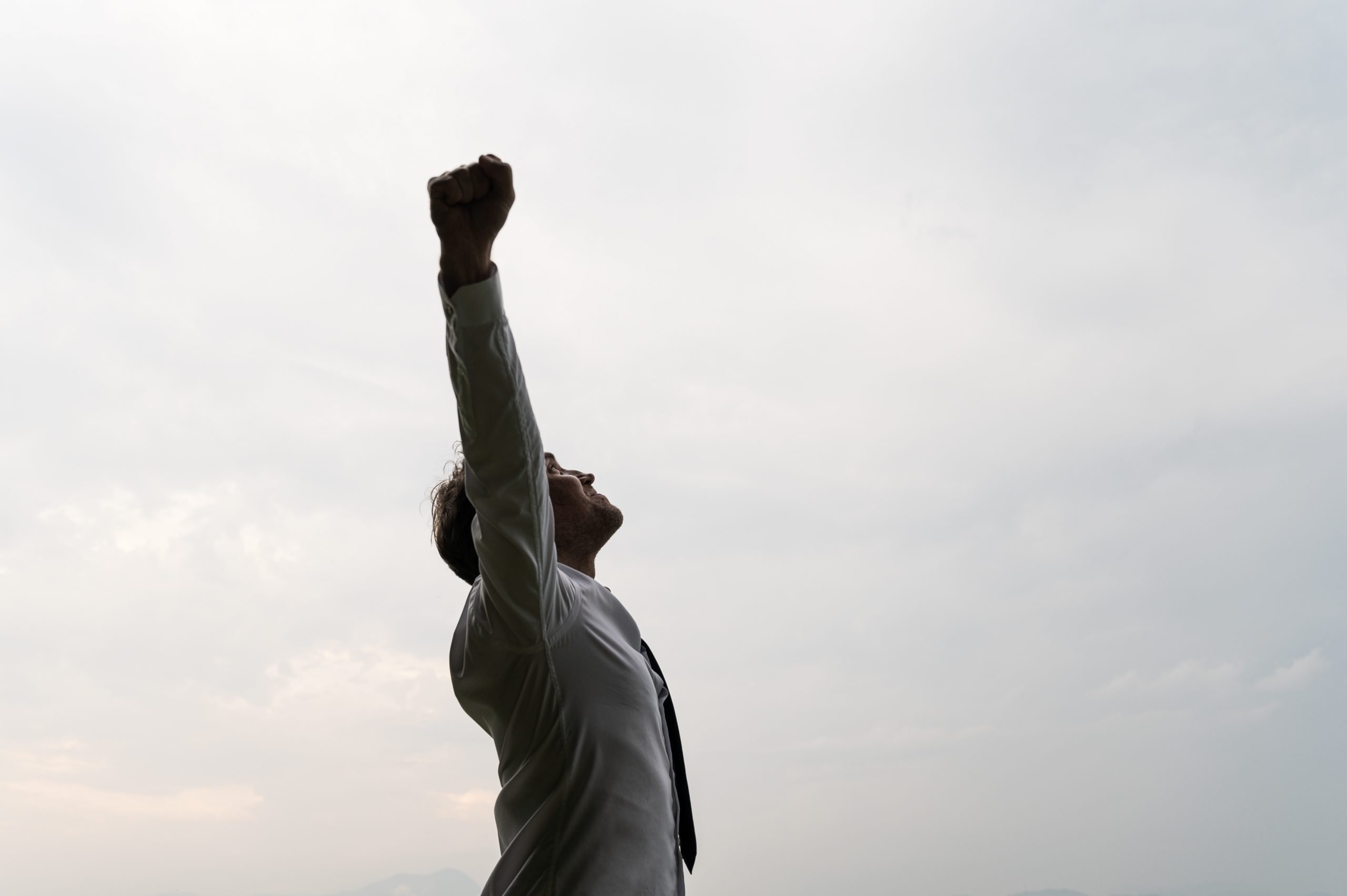 Profile view of successful young businessman standing under cloudy grey sky celebrating success with his arms raised high.