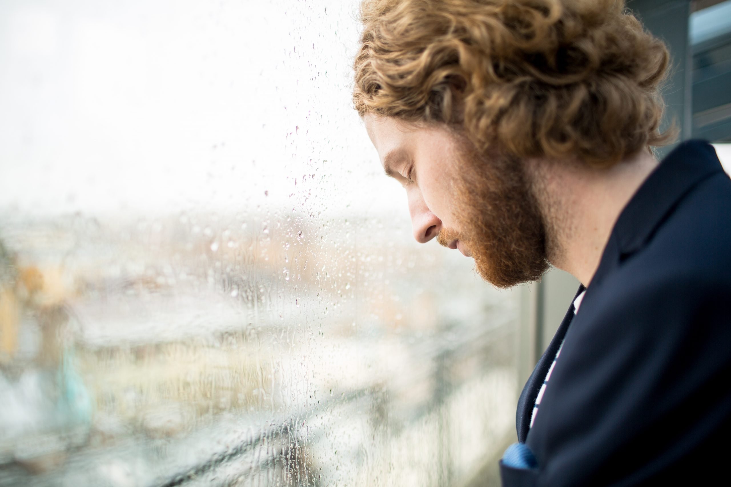 Tired young businessman with his eyes closed touching wet office window by his forehead