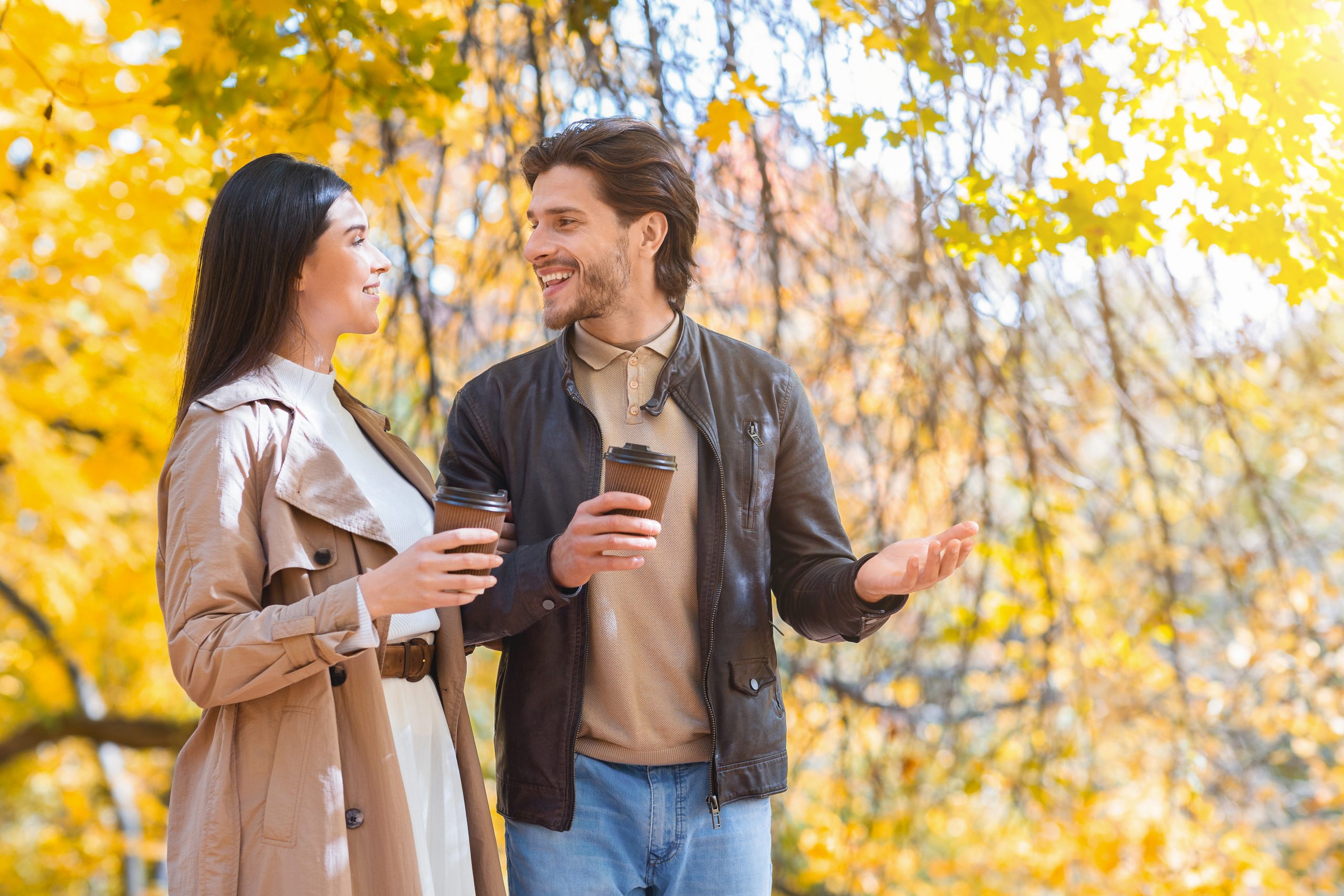 Happily married young couple with cups of coffee walking by autumn forest, having conversation, copy space