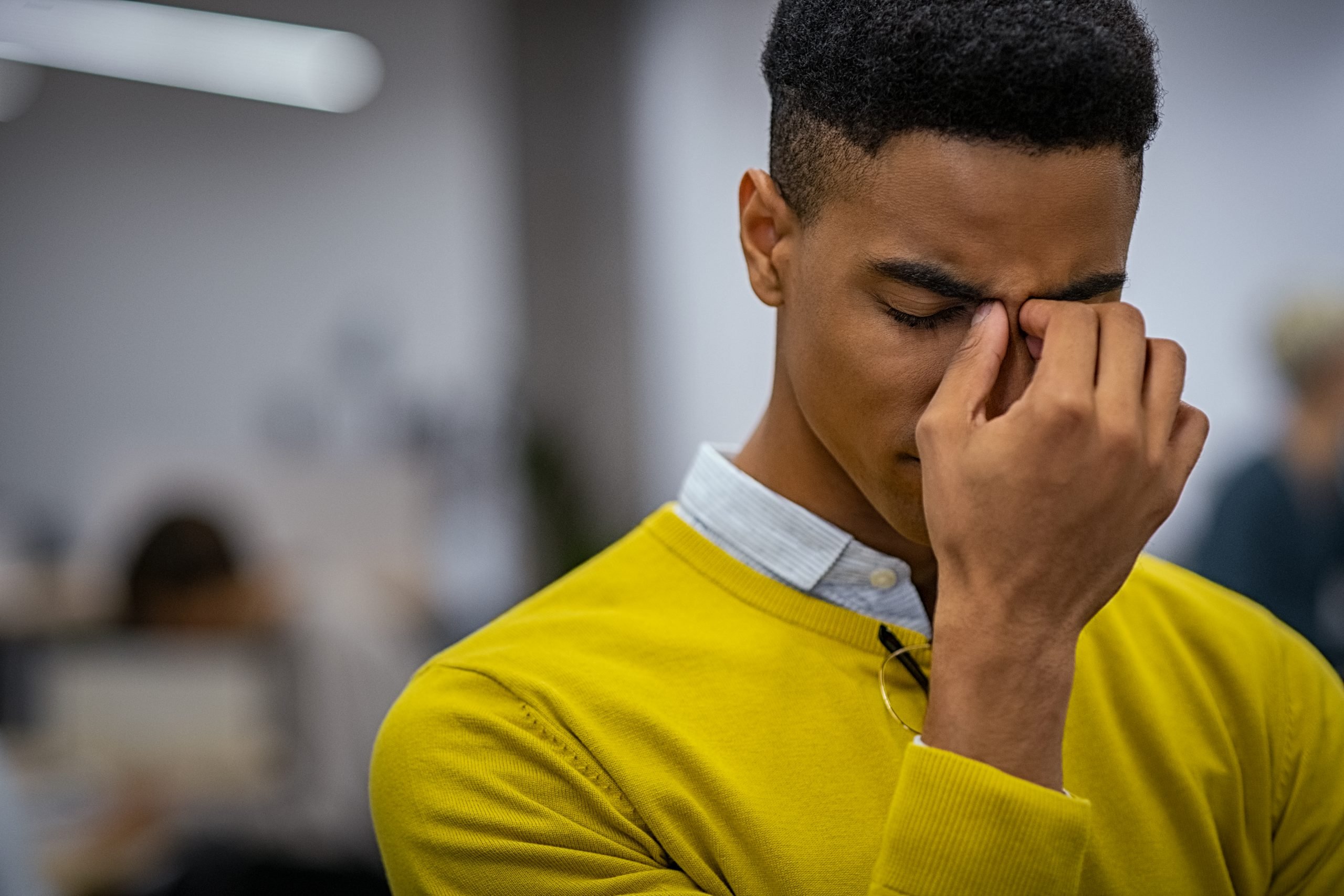 Portrait of an upset young businessman standing in creative office. Black depressed business man with a terrible headache. Stressed african man feeling strain in eyes after working for long hours on computer: overwork, burnout and workload concept.