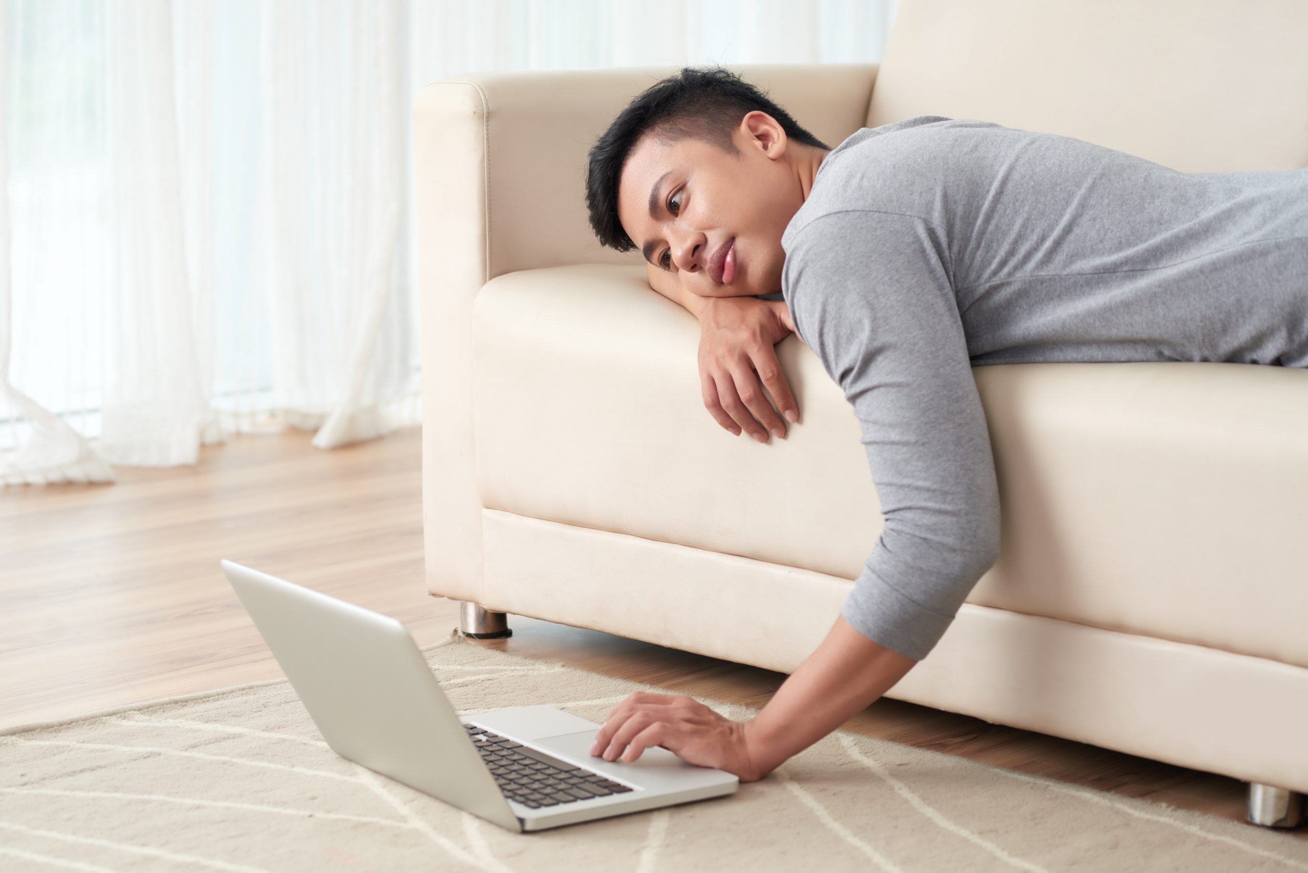 Lazy yuong Vietnamese man lying on sofa and watching something on laptop