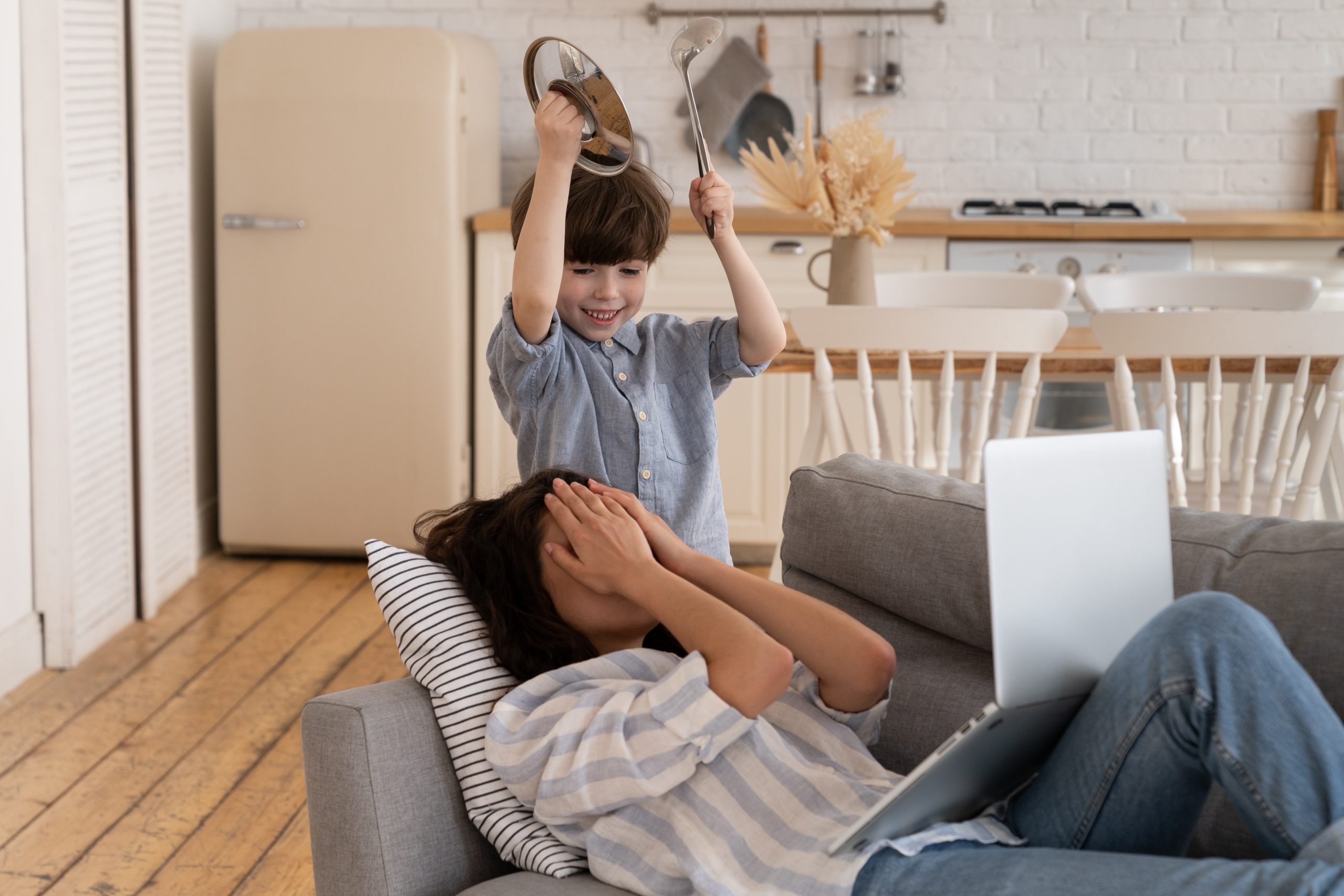 Depressed unhappy mom cover face with hand to protect from noisy son bad behavior. Young mother entrepreneur or freelancer tired of work from home with little kid. Desperate mommy suffer from headache