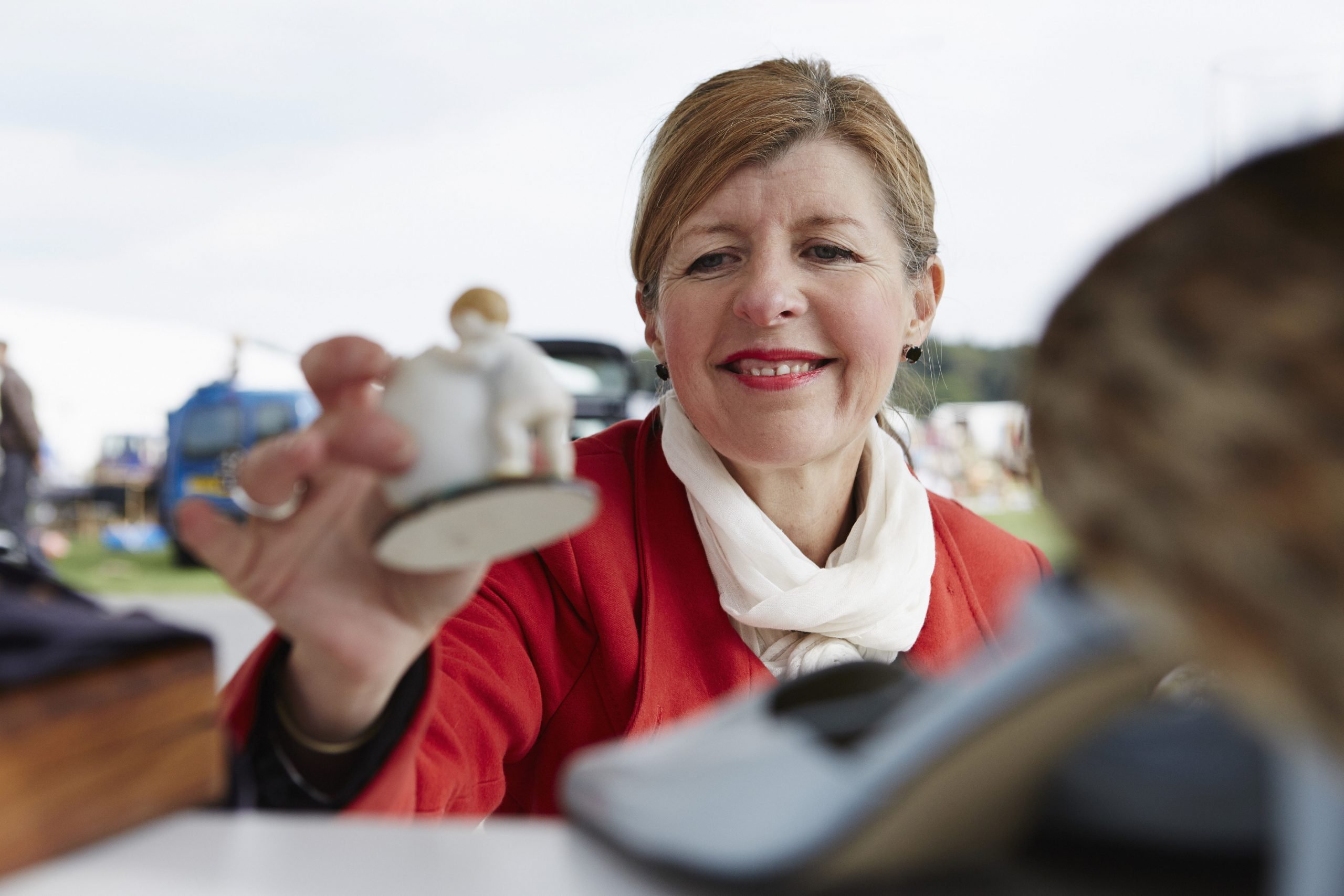 A mature woman in a red coat holding a vintage porcelain figurine at a flea market.