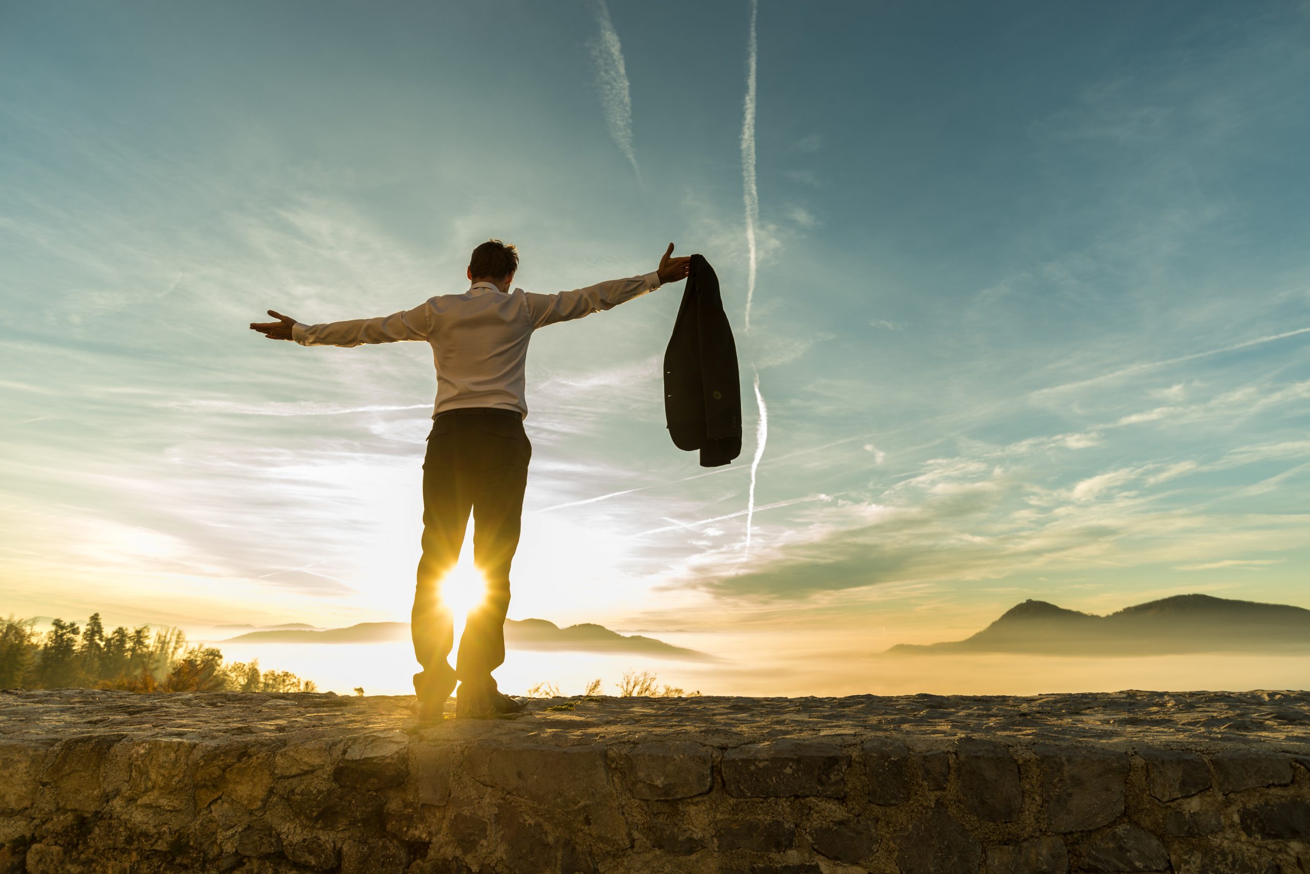 Rear view of successful businessman rejoicing at sunrise standing against the bright glow of the sun with outstretched arms.