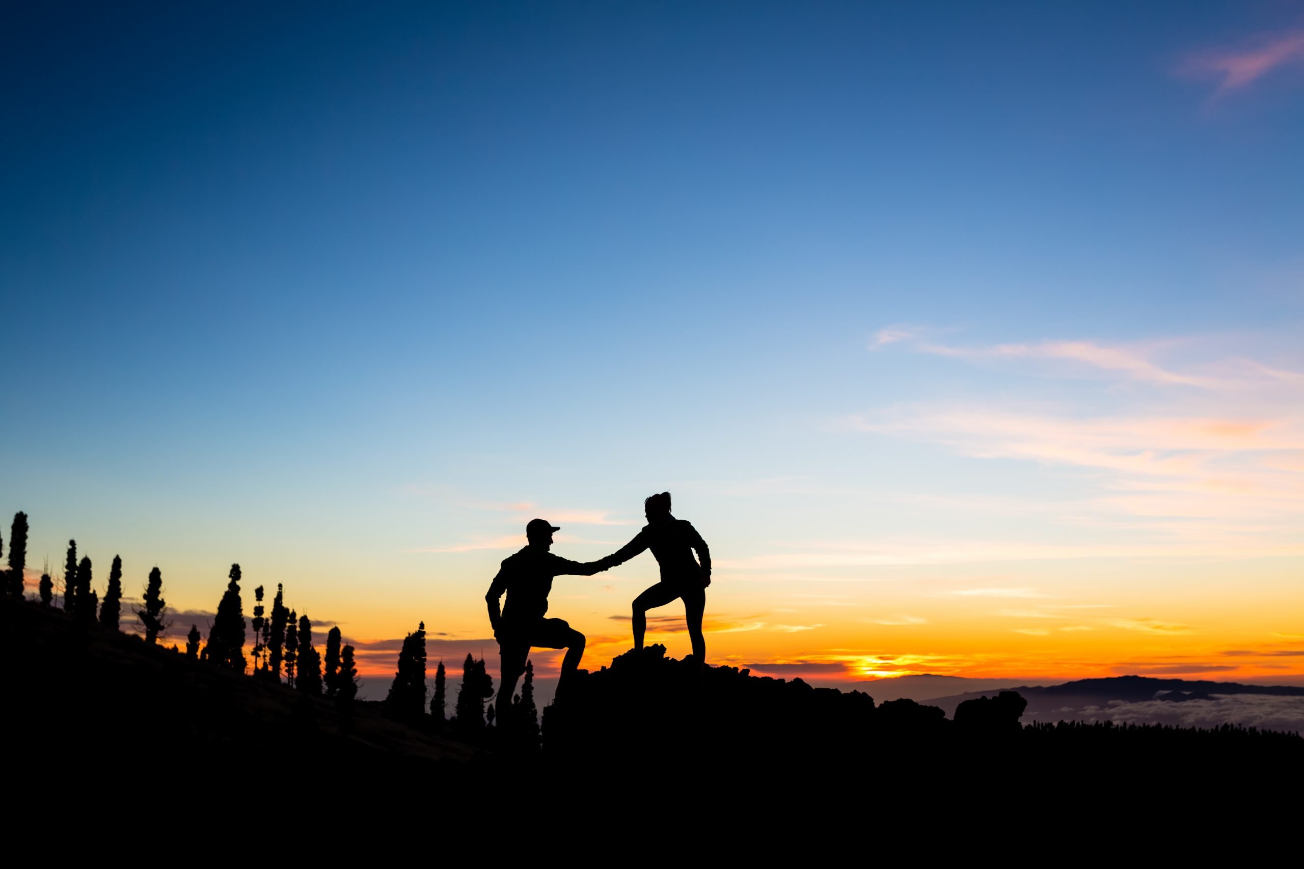 Teamwork couple with helping hand and trust. Silhouette of team help over mountains sunset. Man and woman hikers or climbers reaching goals in inspirational landscape on Tenerife Canary Islands.