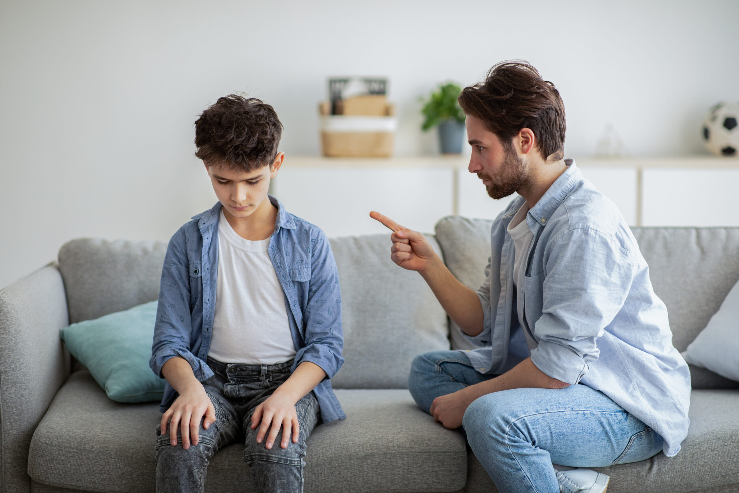 Family discipline concept. Grumpy dad scolding his offended son, teaching his kid tolerance, respect, well-behaving, sitting together on sofa at home interior