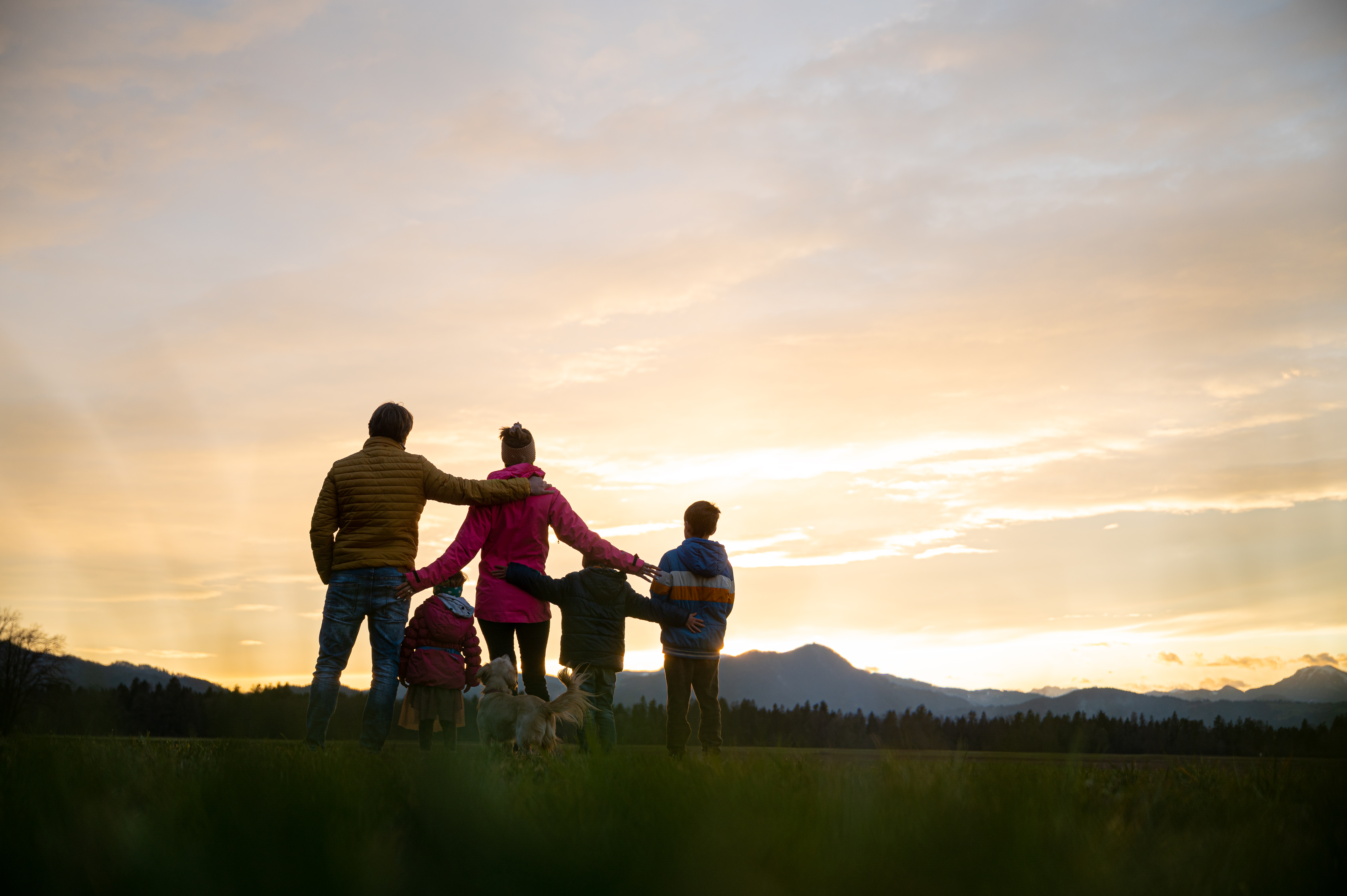 View from behind of family of five and a dog standing outside at dusk with teir arms around each other.