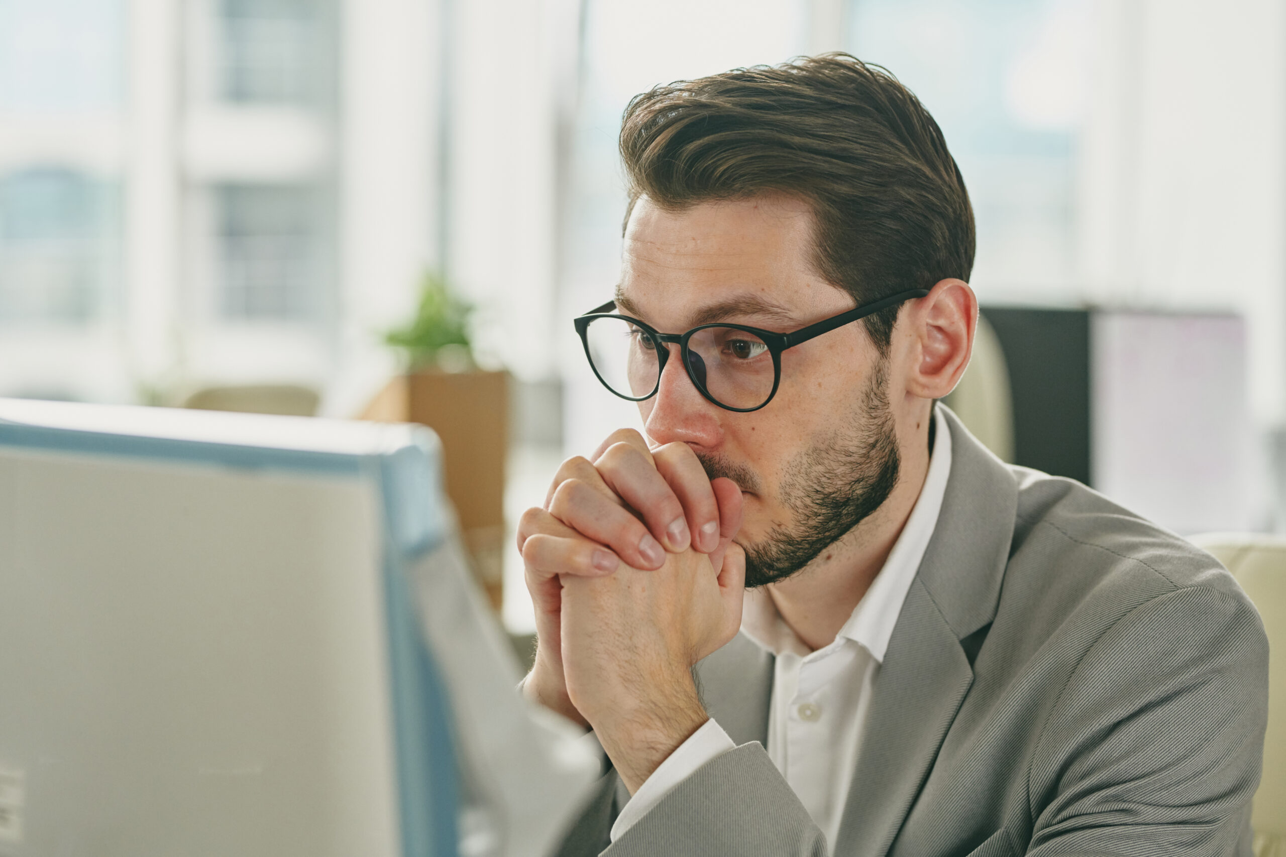 Young pensive businessman in eyeglasses cannot understand information on computer screen while sitting by workplace in office