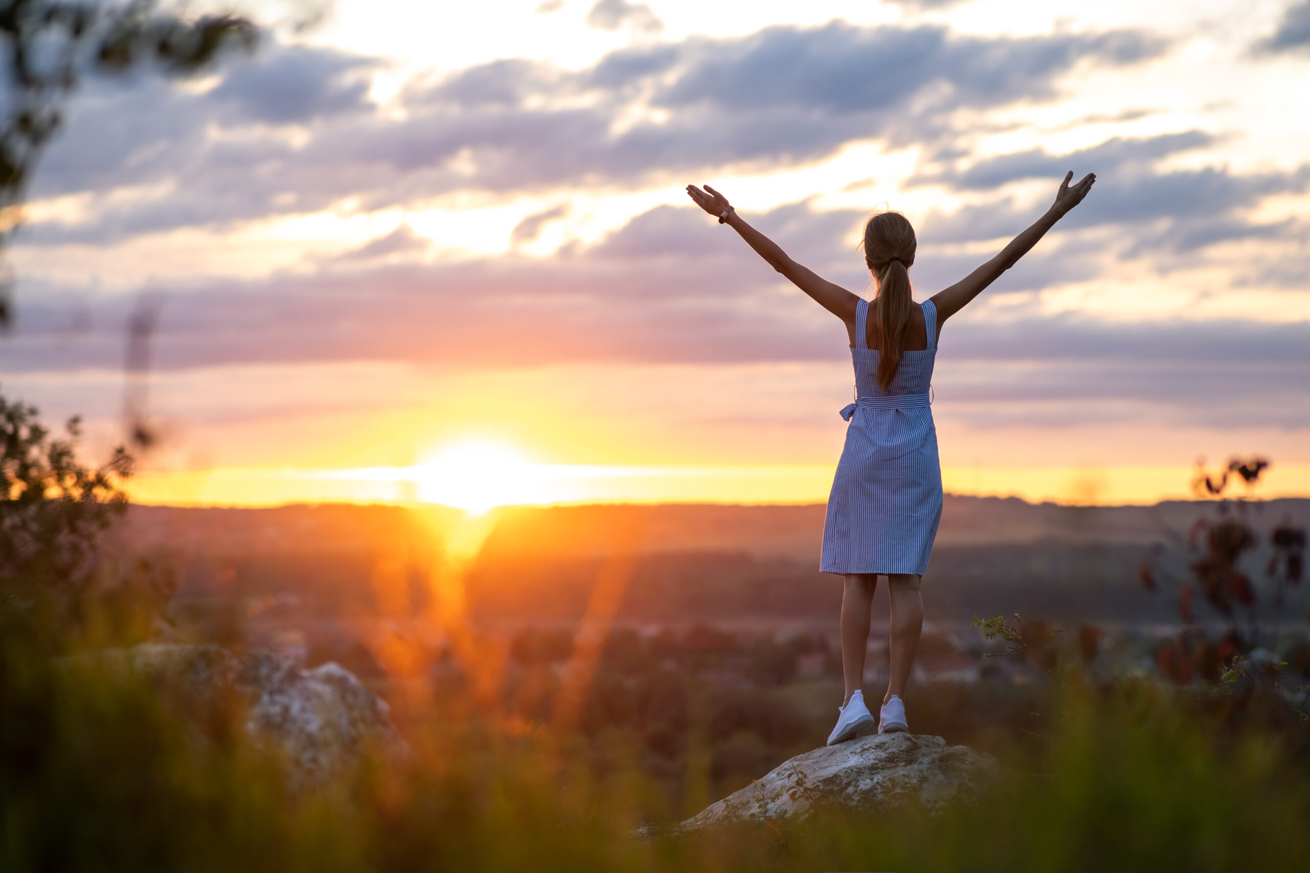 Dark silhouette of a young woman standing with raised up hands on a stone enjoying sunset view outdoors in summer.