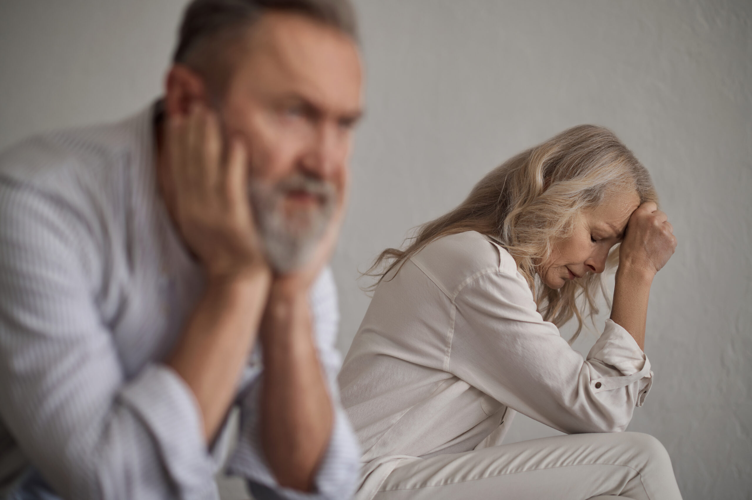 Frustrated blonde lady and a silent gray-haired male sitting apart from each other on the bed