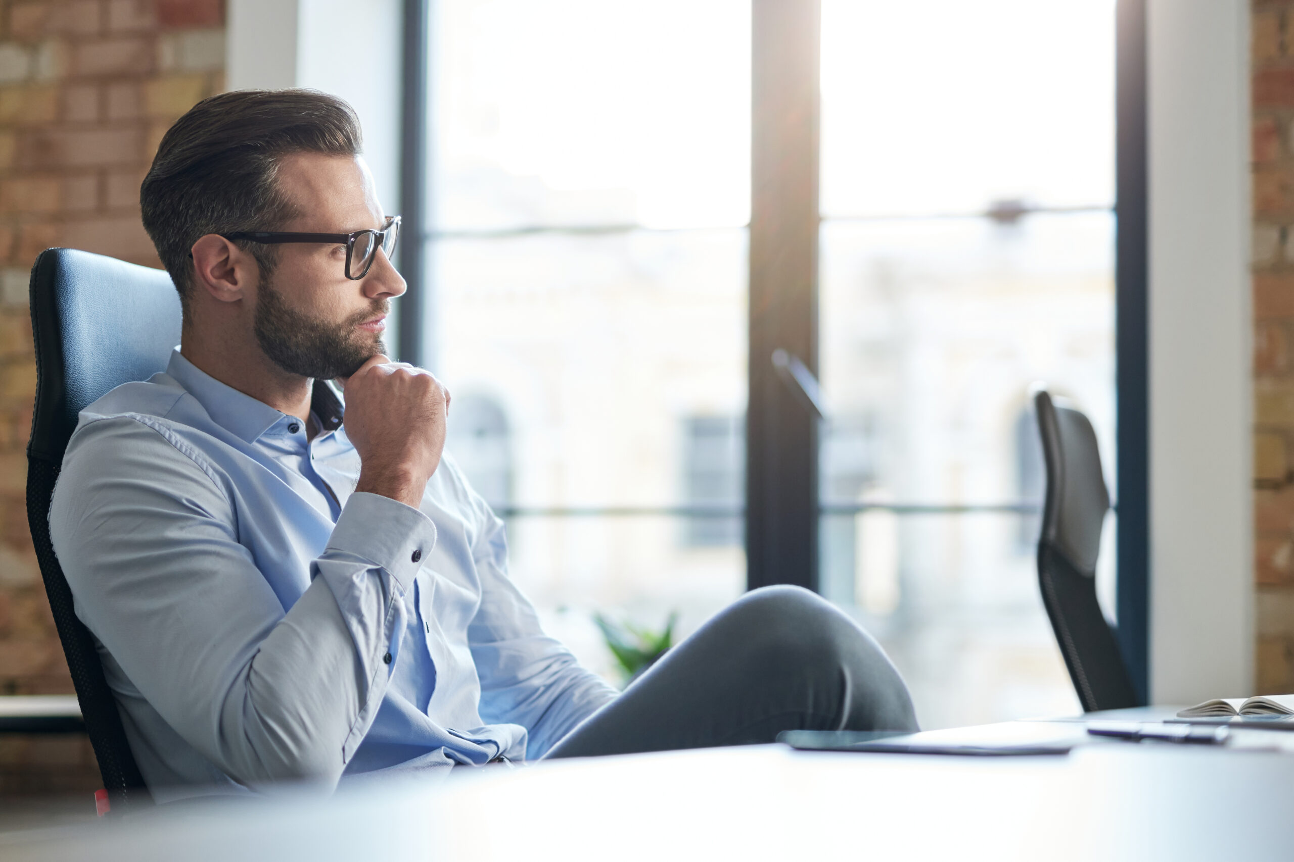 Calm handsome Caucasian man looking away while sitting in the office at daytime and touching his chin