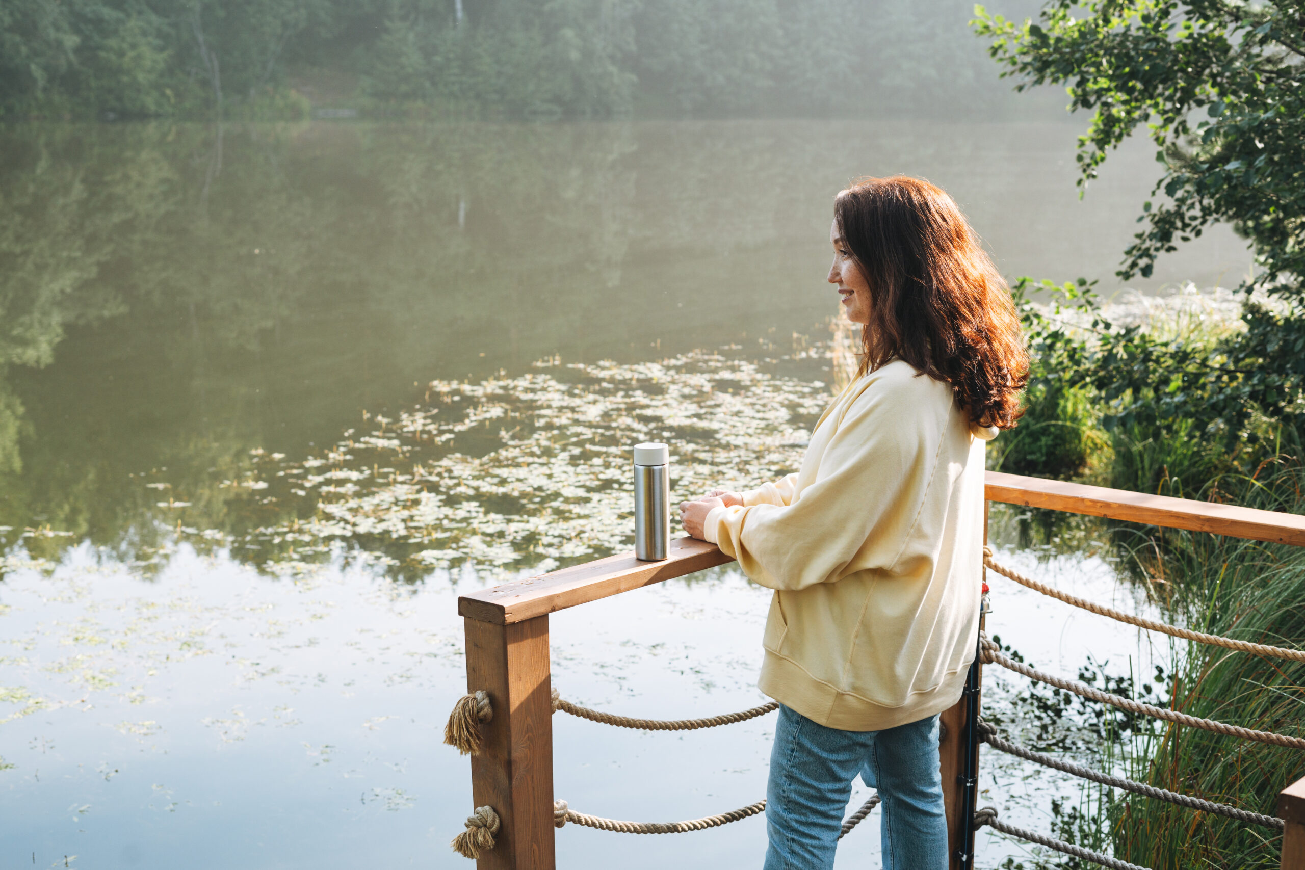 Brunette woman in yellow hoodie with thermo mug relaxing near lake in early morning