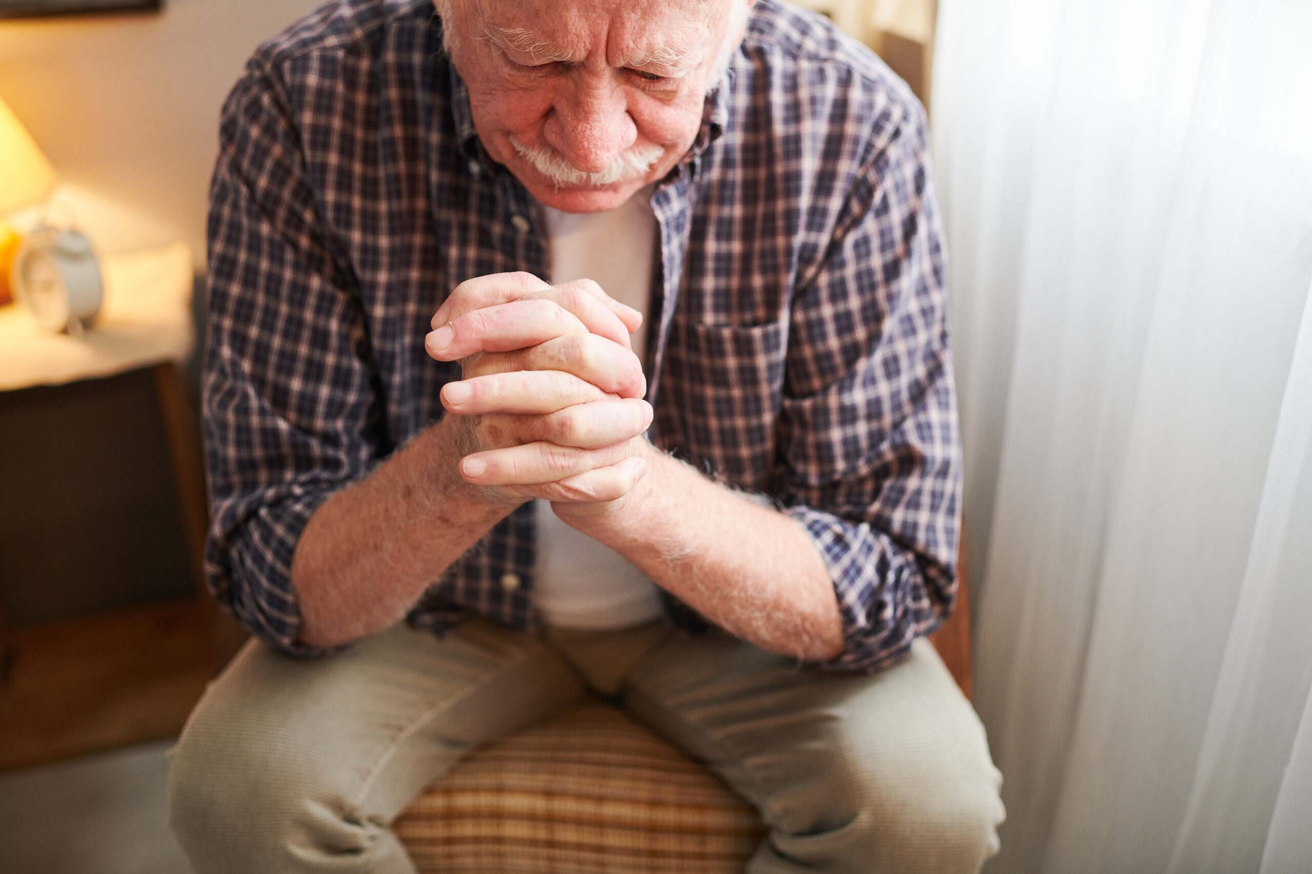 Elderly man with his fingers crossed by his face sitting on chair and praying while resting at home