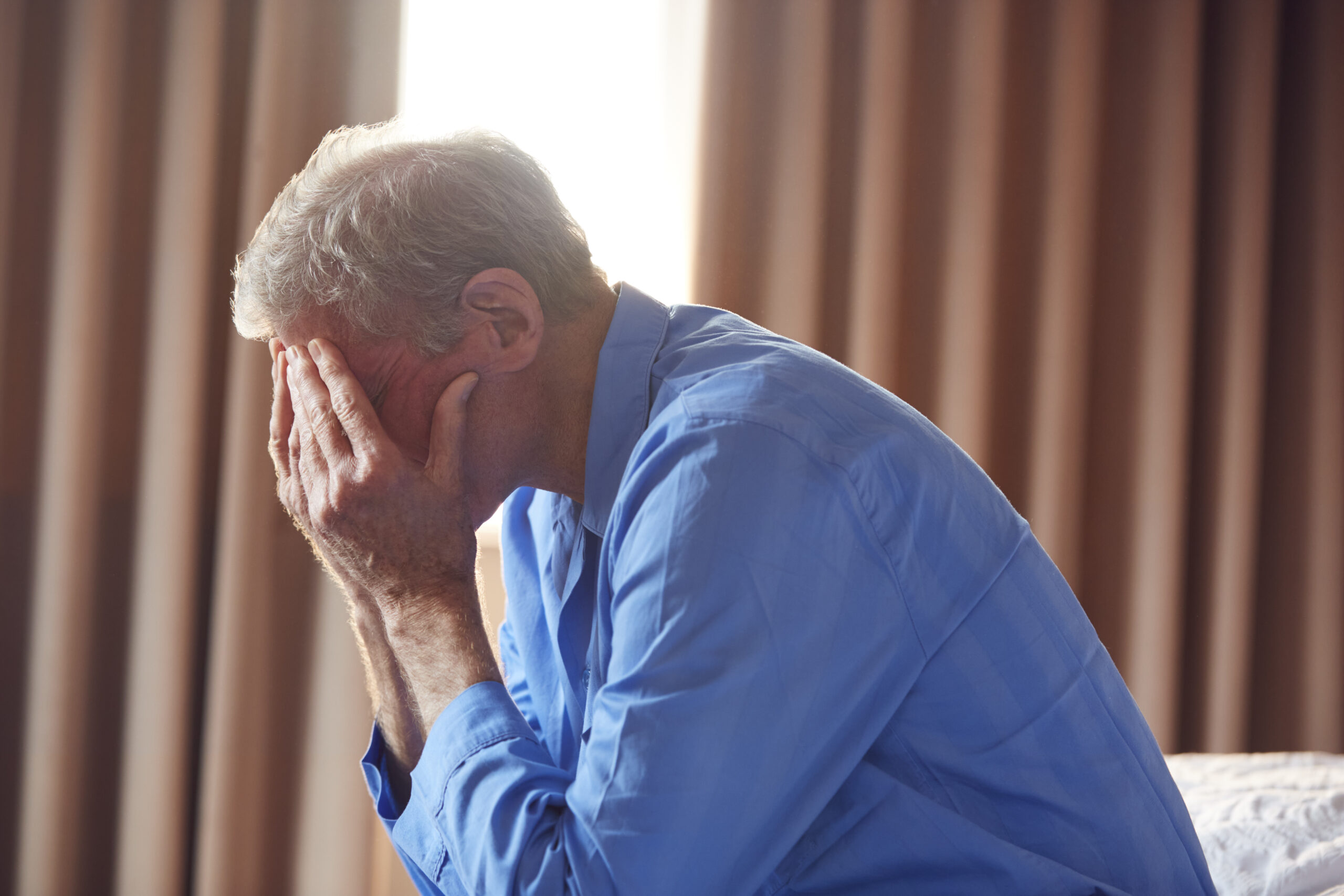 Unhappy And Depressed Senior Man With Head In Hands Sitting On Edge Of Bed At Home