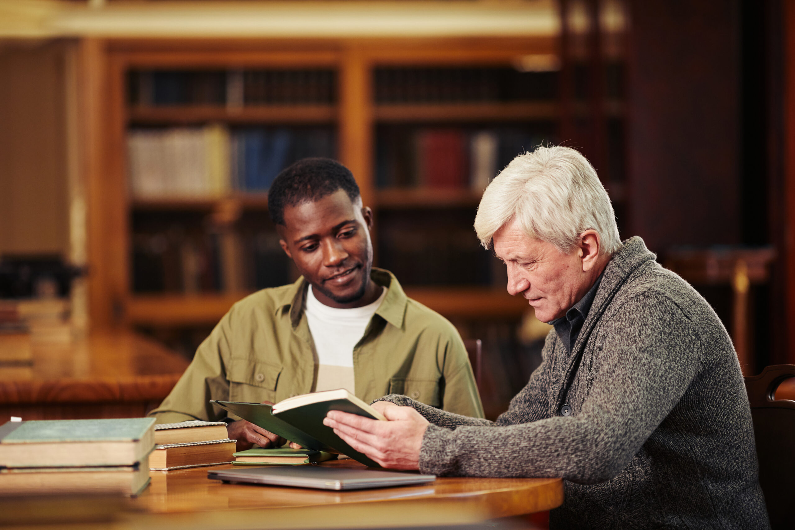 Portrait of male senior student reading book in library while preparing for exam with African-American friend