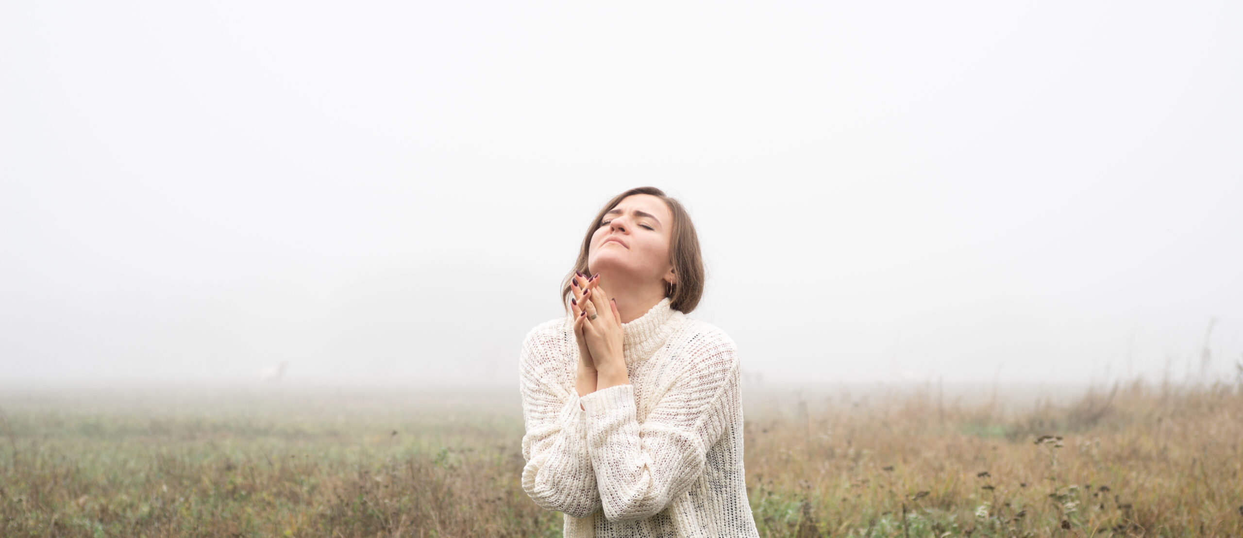 Girl closed her eyes, praying in a field during beautiful fog. Hands folded in prayer concept for faith, spirituality and religion. Peace, hope, dreams concept