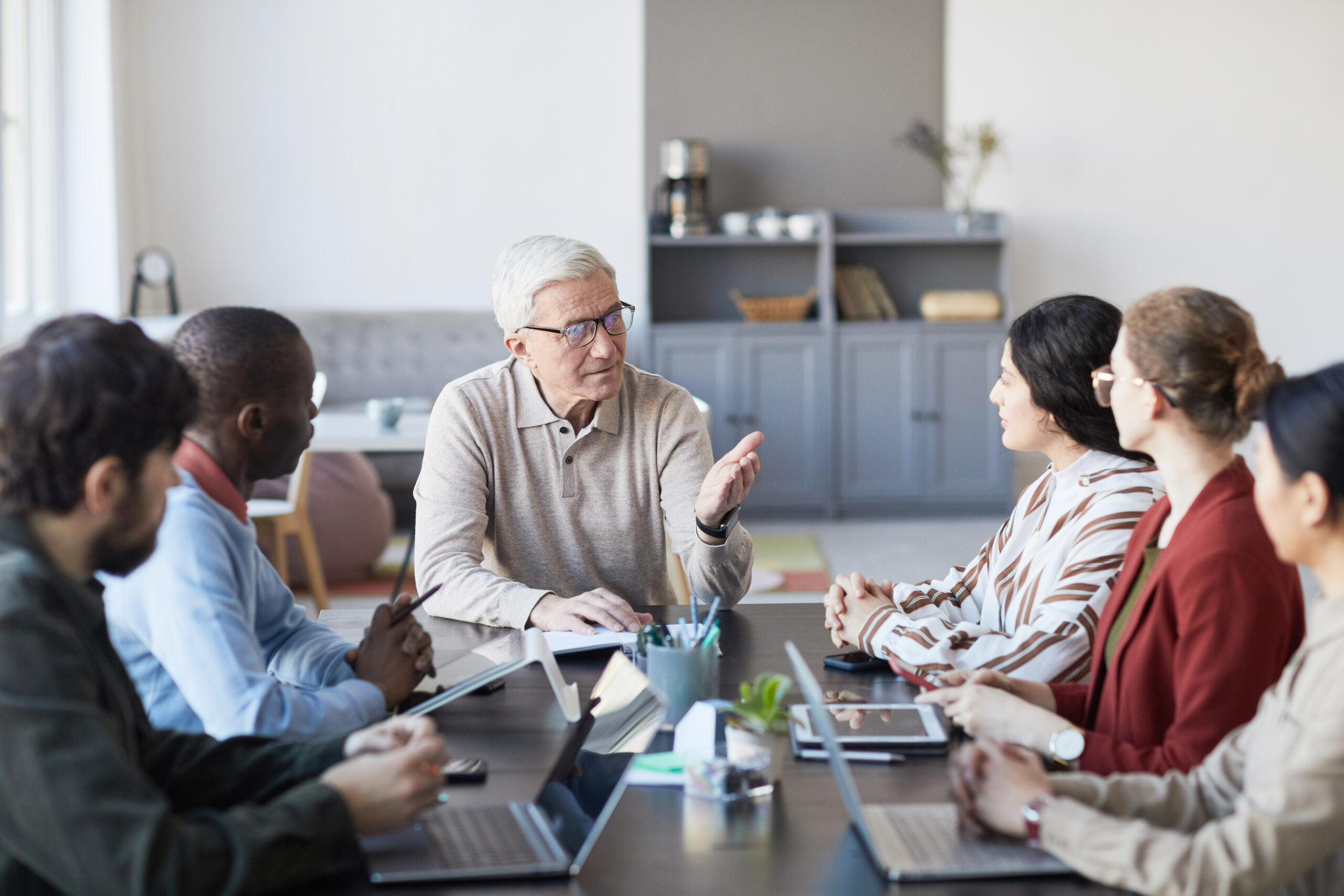 Portrait of modern senior businessman talking to group of people during meeting at table in office