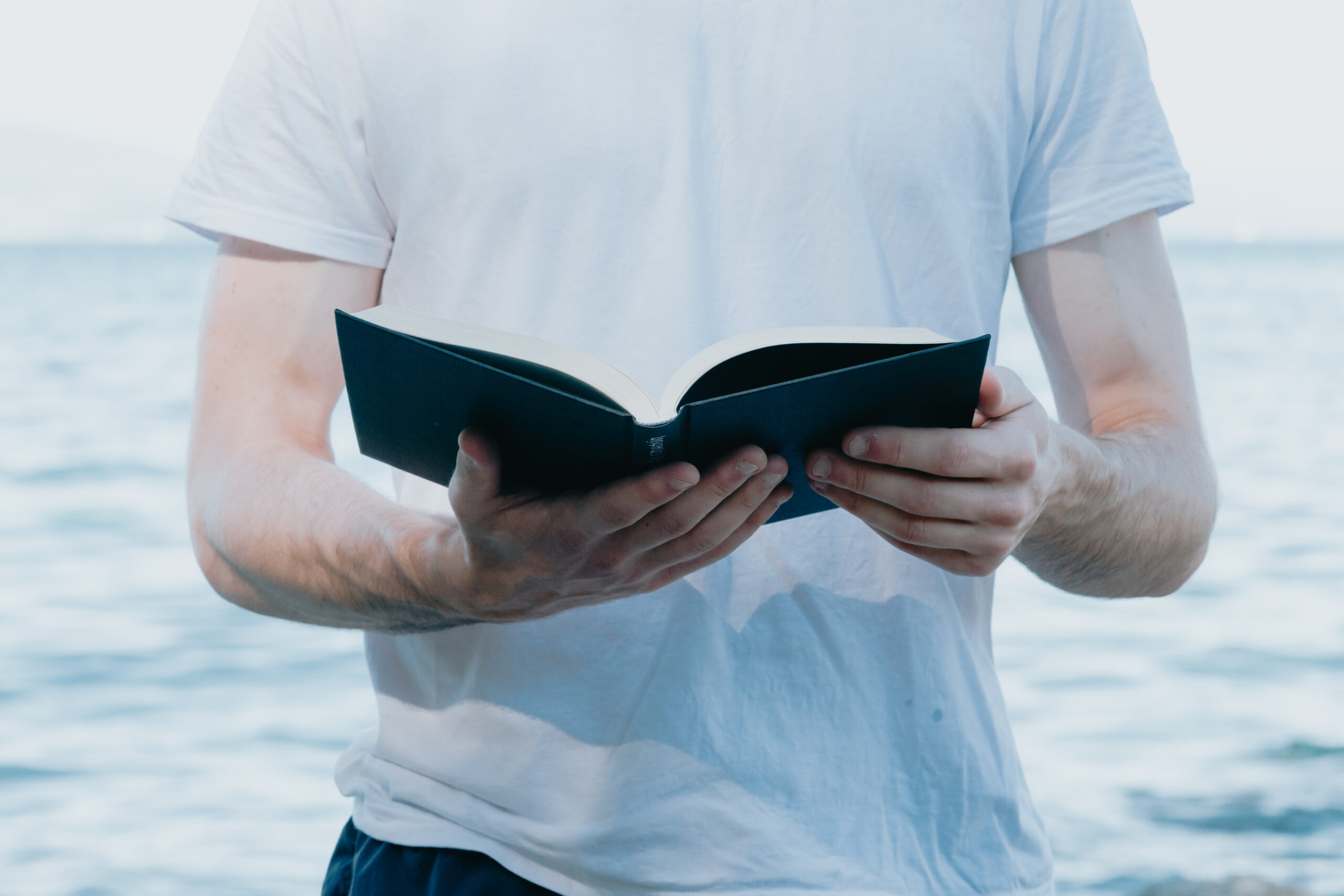 Close up of a man holding a book while reading, religious and study concepts, beach and bright day