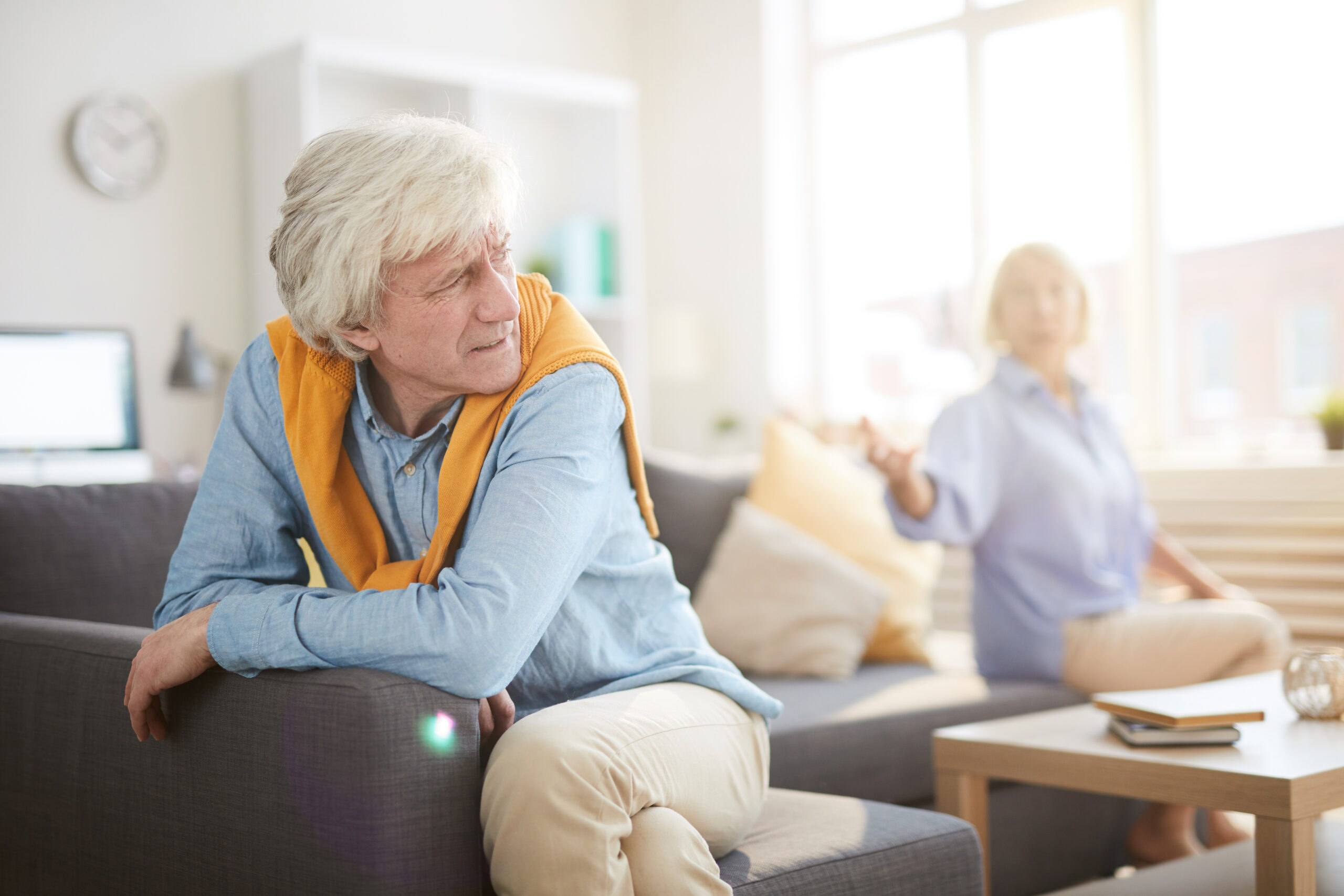 Portrait of senior couple fighting sitting on opposite sides of sofa at home, focus on angry husband in foreground, copy space