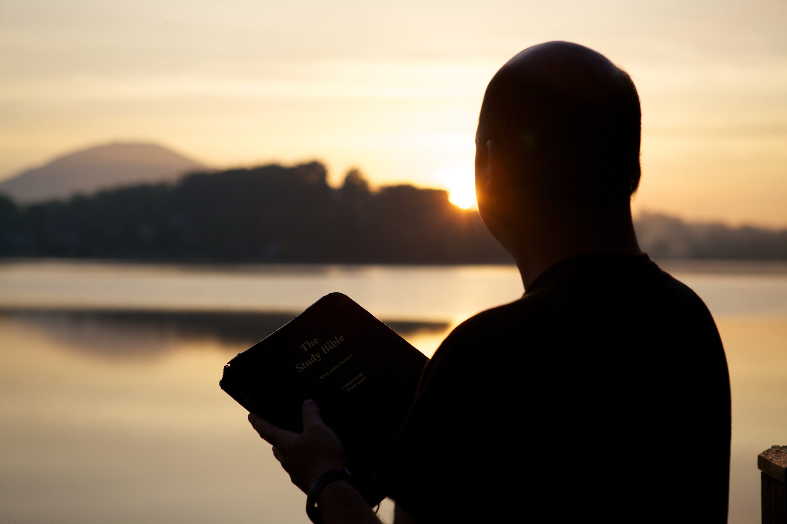 A silhouette of a man holding the bible surrounded by hills and the sea during the sunset