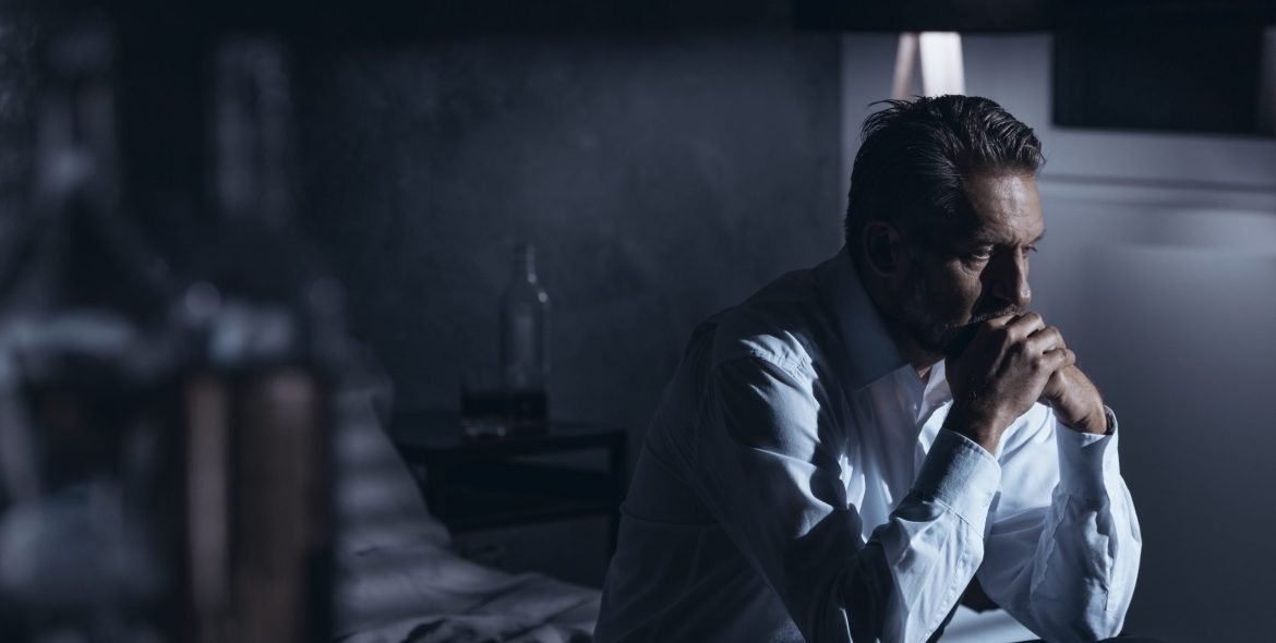 Portrait of a lonely mature man with depression sitting on a bed in a gray room with bottles of alcohol standing around