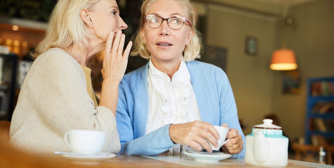 One of pretty aged females telling secret to her friend by cup of tea in cafe