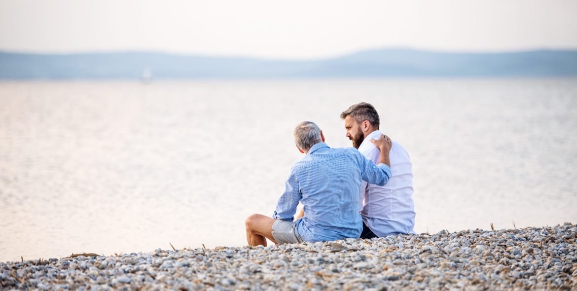 Rear view of senior father and mature son sitting by the lake, talking. Copy space.