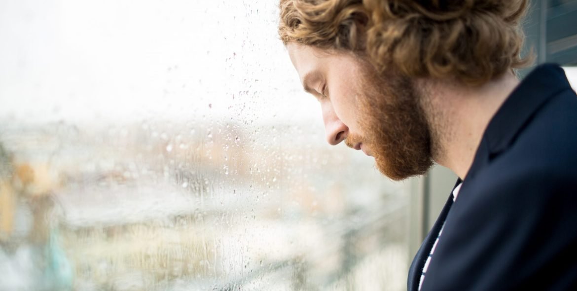 Tired young businessman with his eyes closed touching wet office window by his forehead