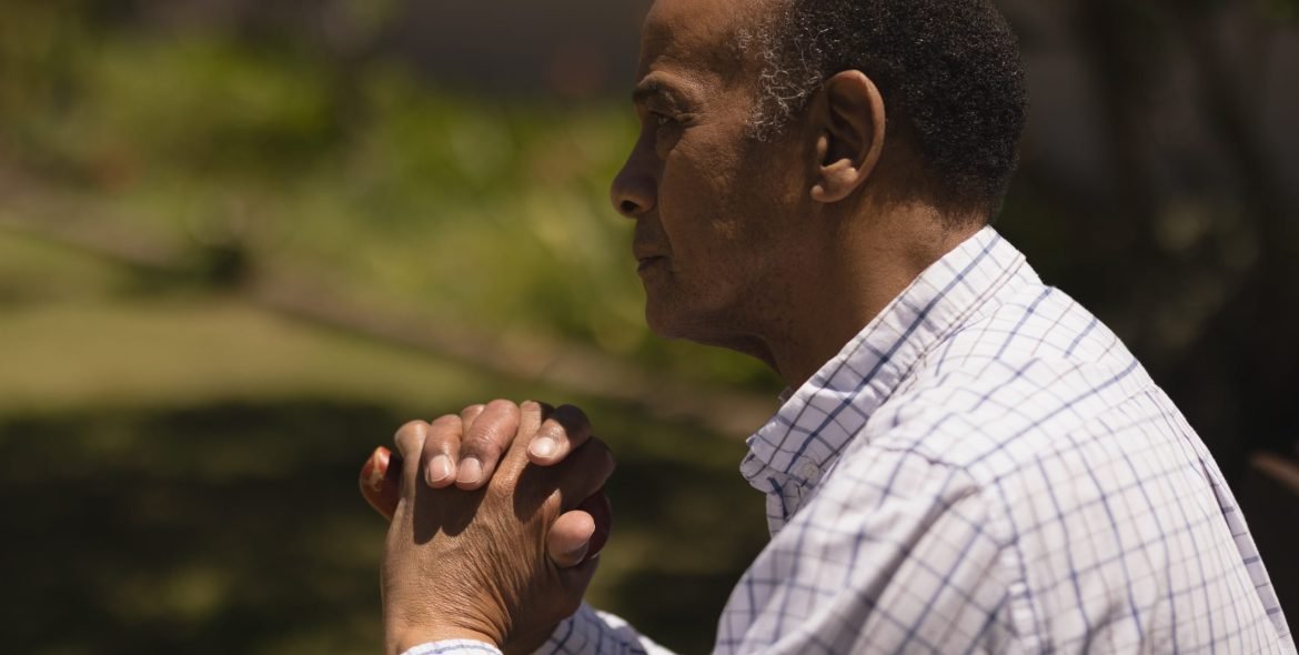 Side view of thoughtful senior man hands leaning on a cane while sitting on bench on sunny day in garden
