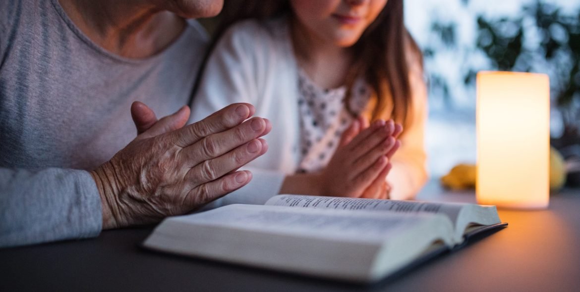 A small girl and her unrecognizable grandmother with bible at home, praying. Family and generations concept.