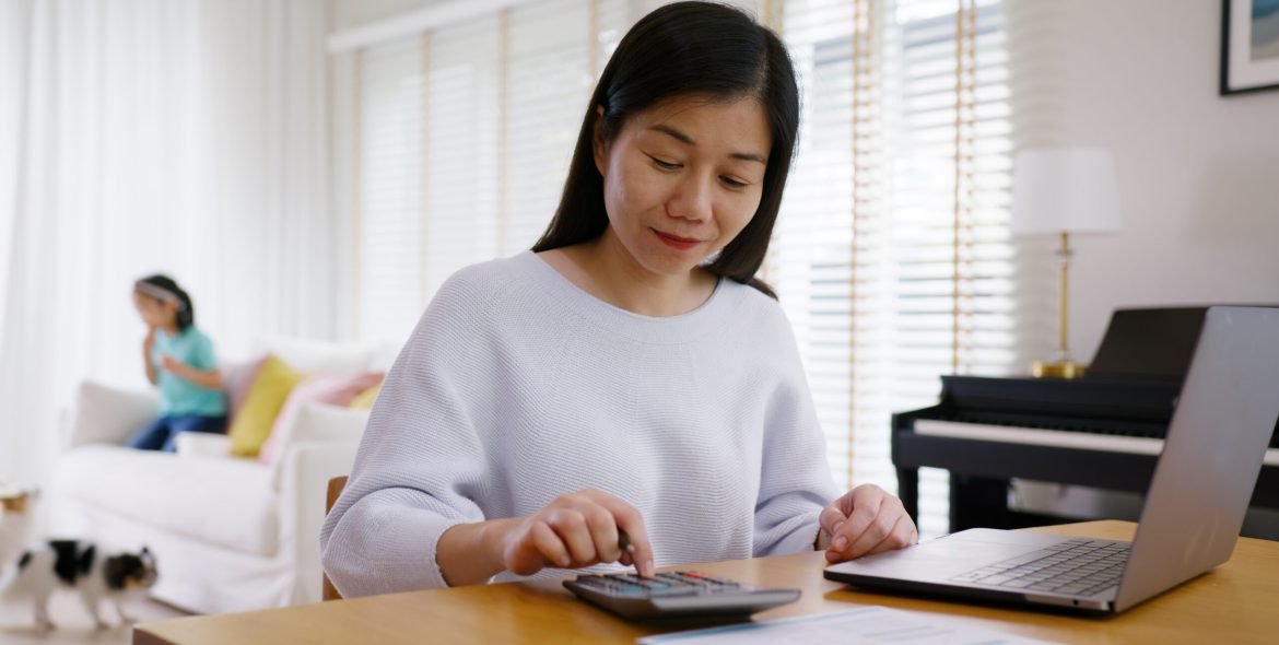 Adult asia people single mom small SME owner support happy sit busy apply child tax credit refund plan form app or pay loan bill on laptop. Little kid girl play at home hybrid job career workforce.