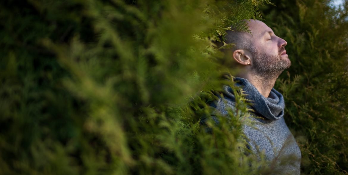 A bald man in a warm sweater is resting, leaning against the trees. Breathe fresh air in nature.