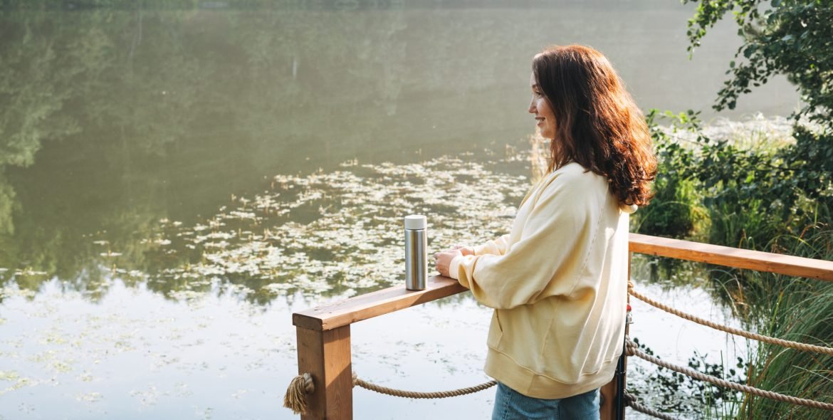Brunette woman in yellow hoodie with thermo mug relaxing near lake in early morning