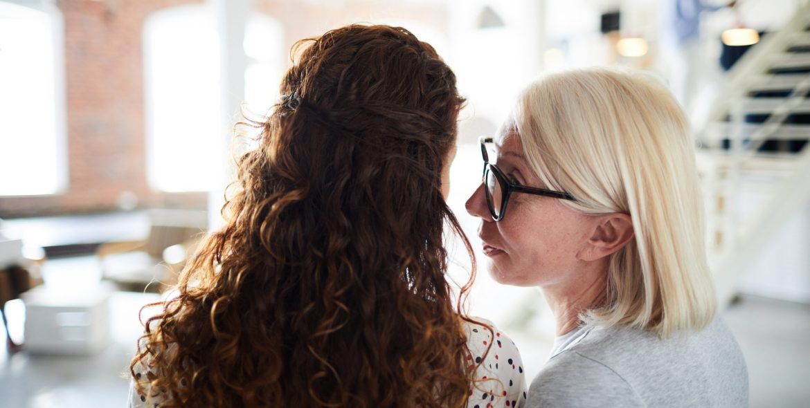 Rear view of mature woman in eyeglasses telling new gossip about colleagues at office