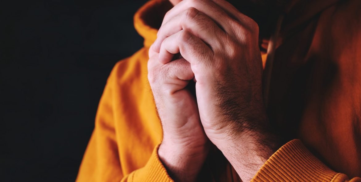 Christian man praying to God in dark room with clasped hands, close up with selective focus