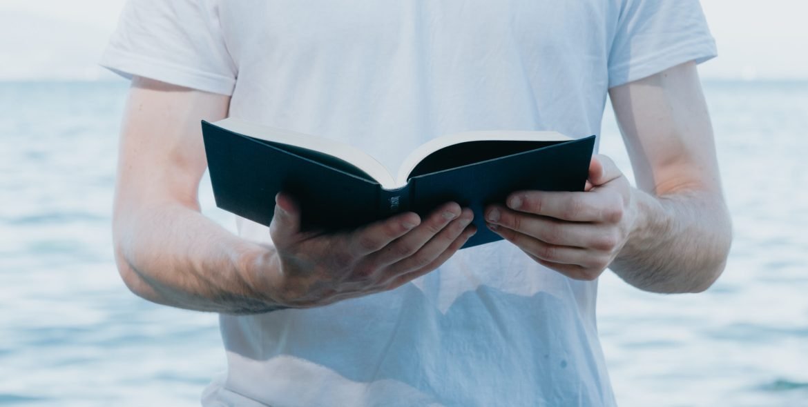 Close up of a man holding a book while reading, religious and study concepts, beach and bright day