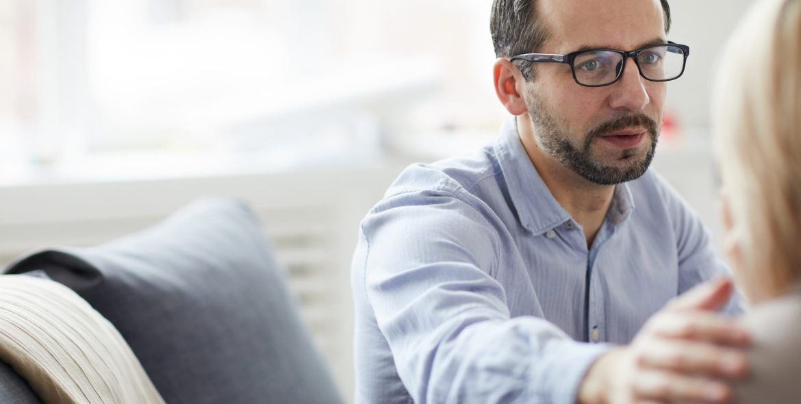 Mature counselor in shirt and eyeglasses touching shoulder of patient while comforting her during session