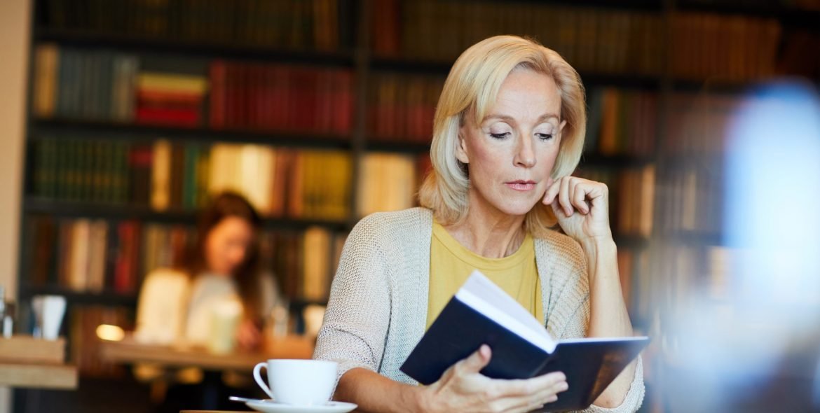 Pensive blonde mature woman sitting by desk in library or cafe and reading book by cup of tea