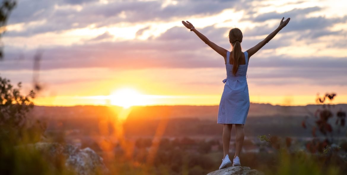 Dark silhouette of a young woman standing with raised up hands on a stone enjoying sunset view outdoors in summer.