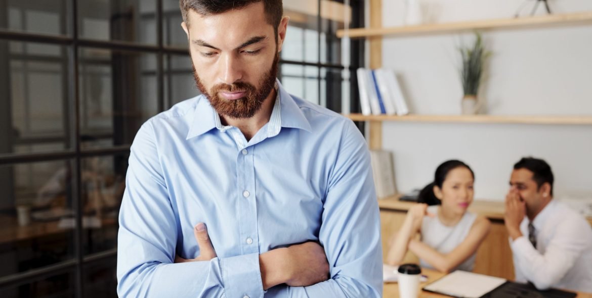 Upset young businessman standing in closed pose, colleagues are gossiping about him in background