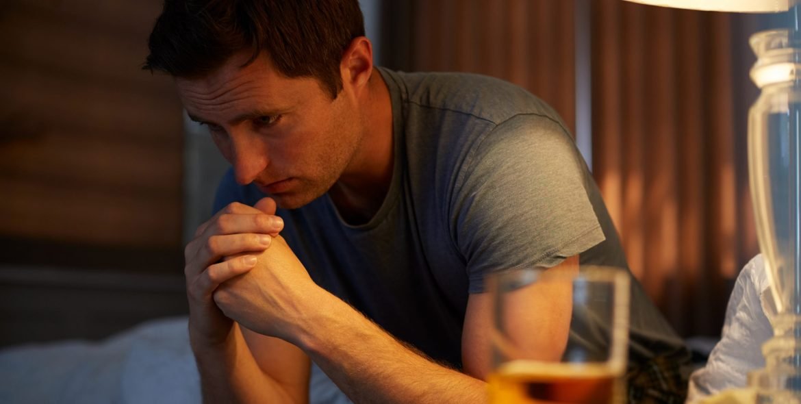 Depressed Man Wearing Pajamas Sitting On Side Of Bed With Glass Of Whisky On Bedside Cabinet