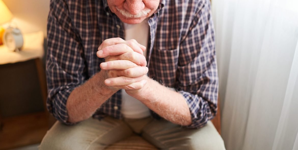 Elderly man with his fingers crossed by his face sitting on chair and praying while resting at home