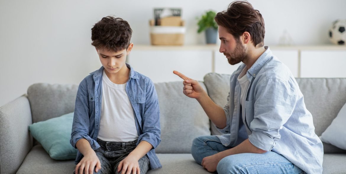 Family discipline concept. Grumpy dad scolding his offended son, teaching his kid tolerance, respect, well-behaving, sitting together on sofa at home interior