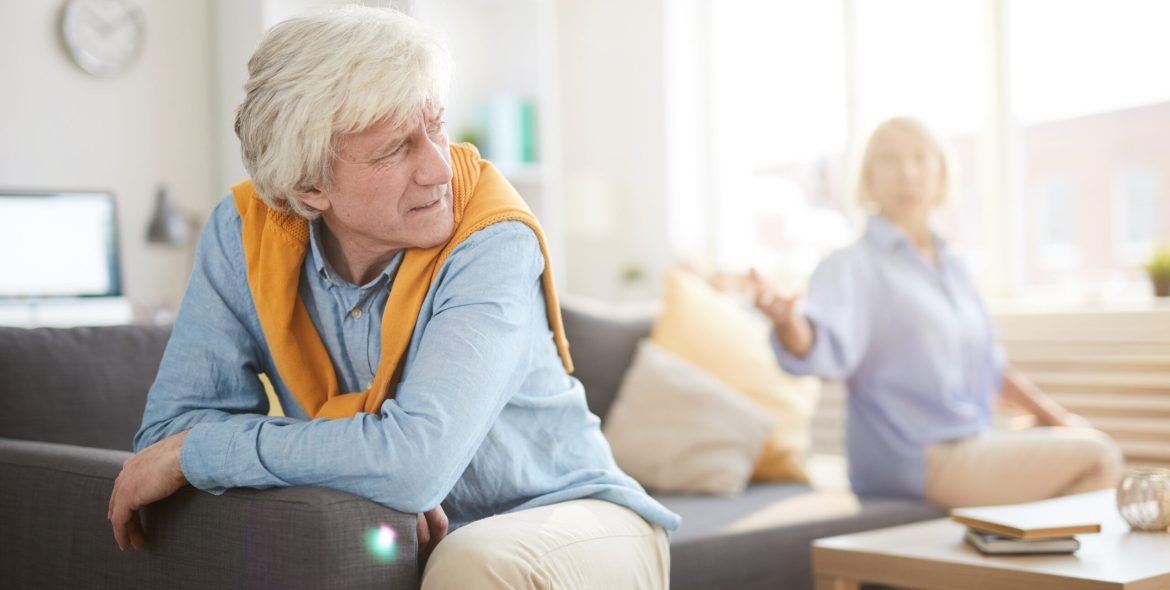 Portrait of senior couple fighting sitting on opposite sides of sofa at home, focus on angry husband in foreground, copy space